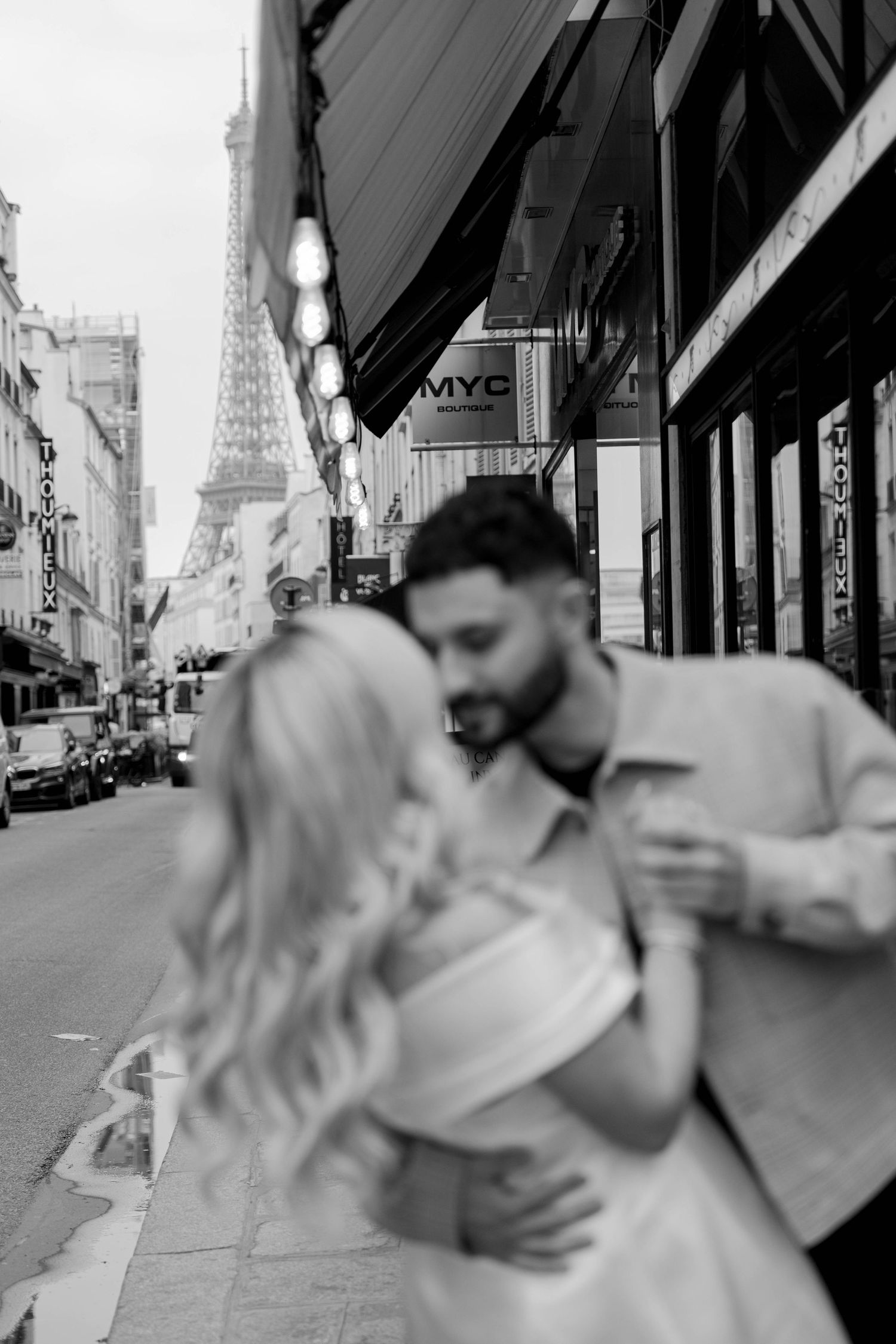 A romantic couple embrace on a Paris street with the iconic Eiffel Tower visible in the background.