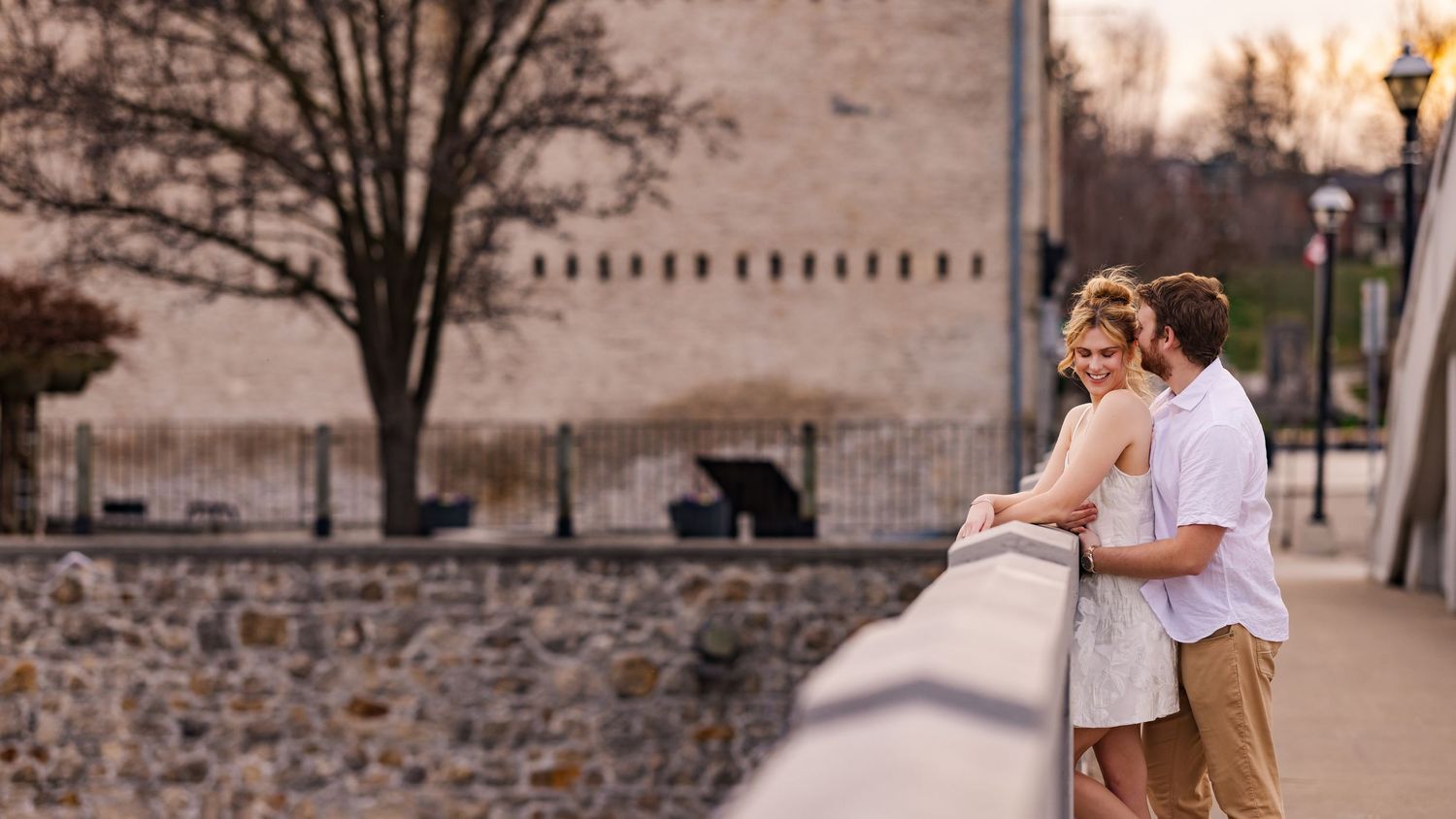 A romantic couple embraces during a sunset photoshoot along a stone wall at a scenic location.