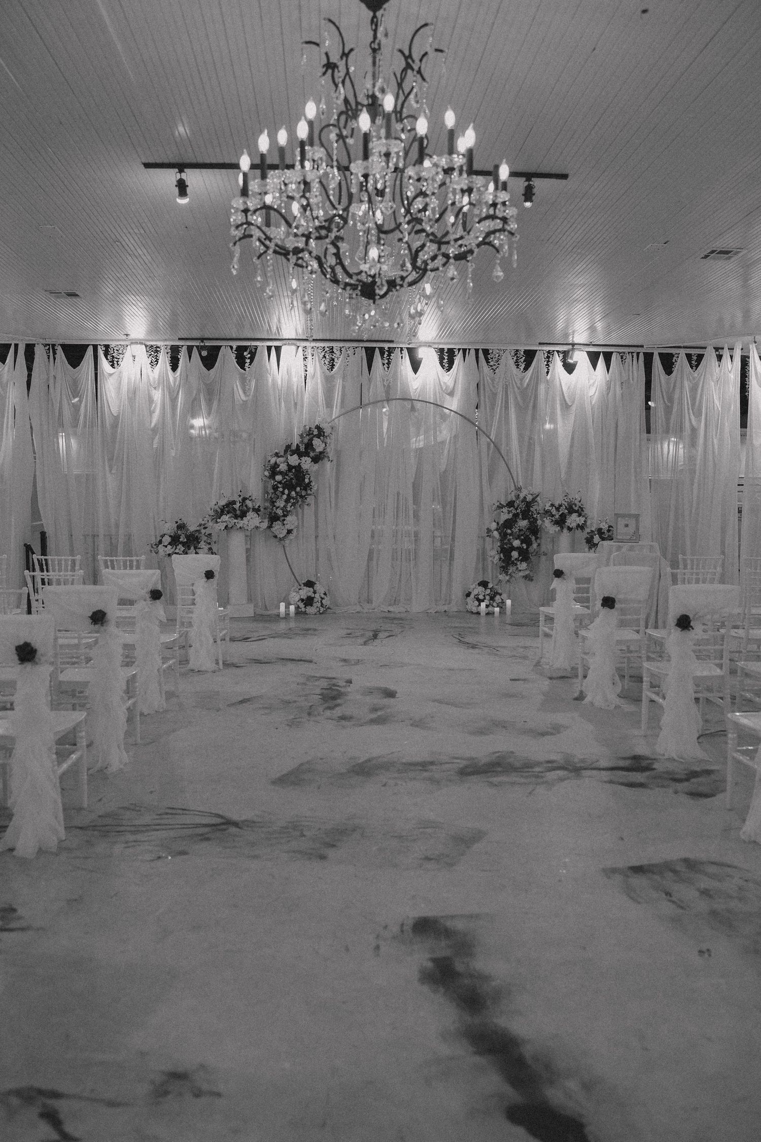 Elegant wedding ceremony setup with crystal chandelier, white drapery and decorative aisle in black and white photography.