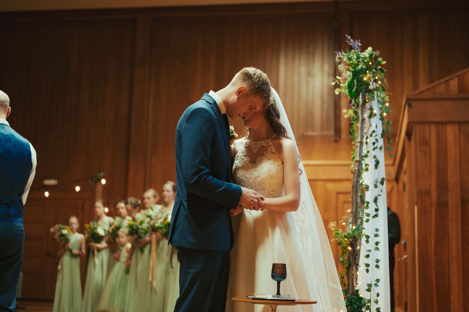 Wedding ceremony guests applaud as a couple walks down the dimly lit aisle with string lights and candles glowing.
