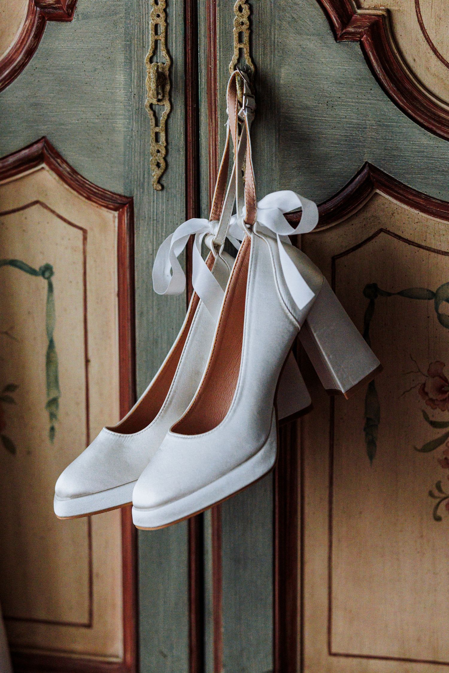 White bridal heels with ribbon bows hanging on an ornate vintage painted wooden armoire door.