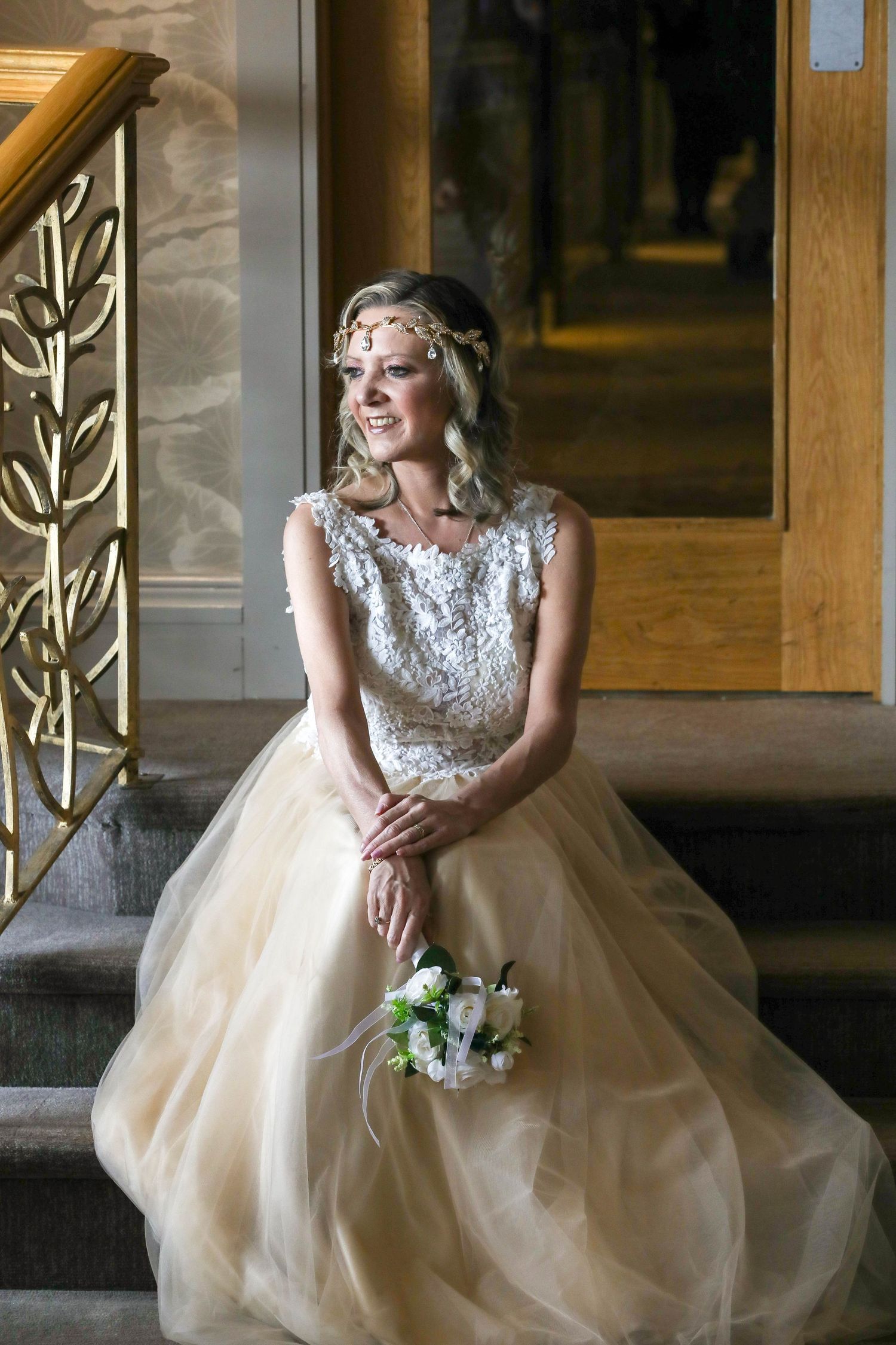 Elegant bride in champagne tulle gown with lace bodice and crystal headpiece sits on stairs holding white bouquet.