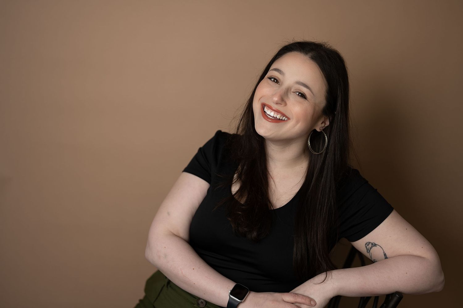 Brunette woman smiling warmly while seated wearing black t-shirt with tattoo in studio portrait.
