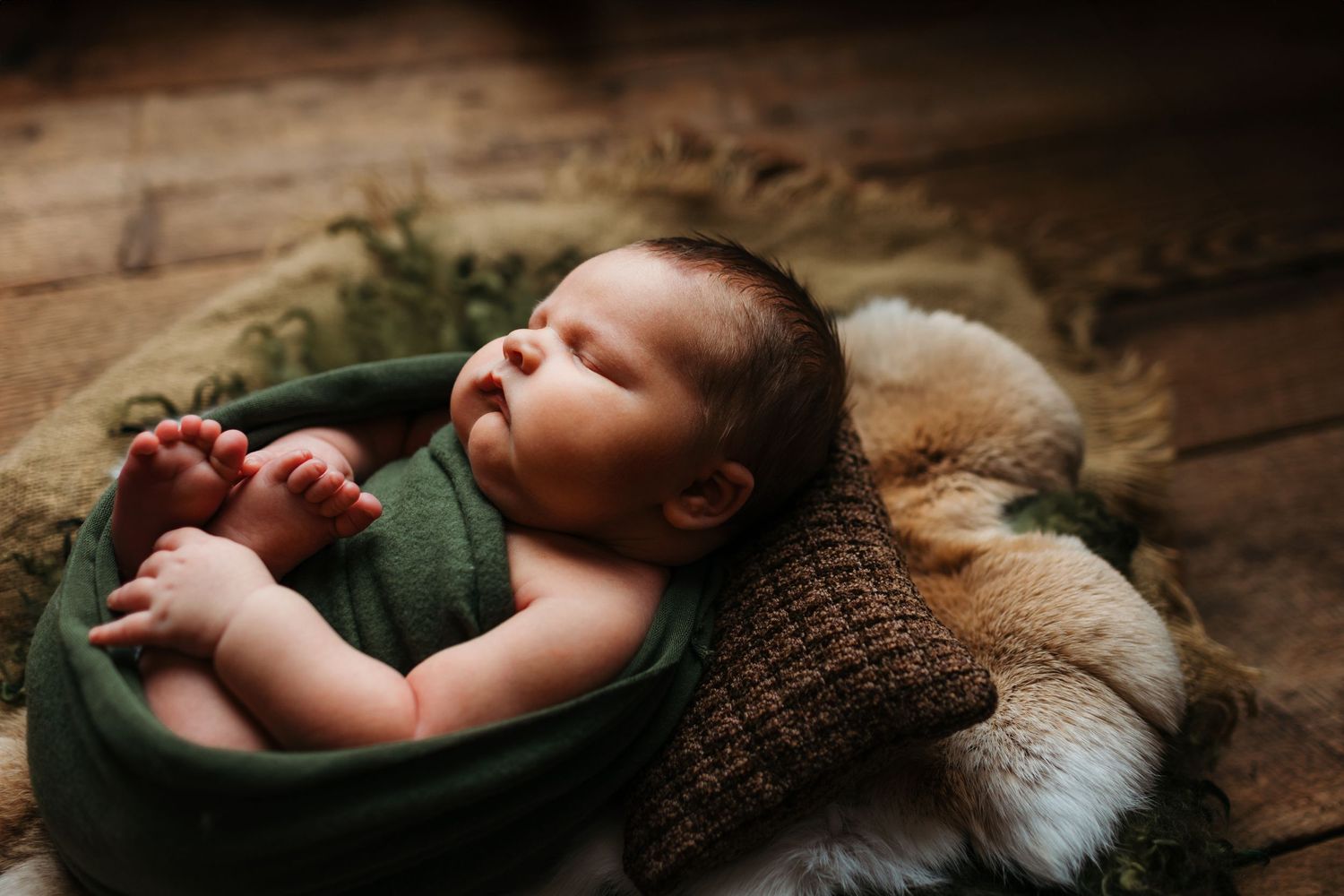Sleeping newborn wrapped in brown knit blanket on textured wooden surface.