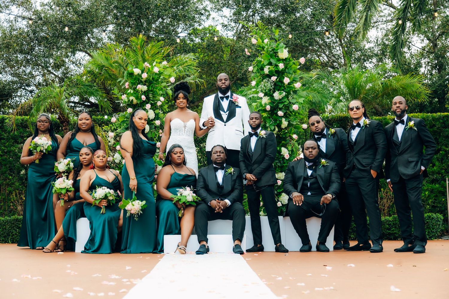 Wedding party in emerald green and black formal attire poses in garden setting.