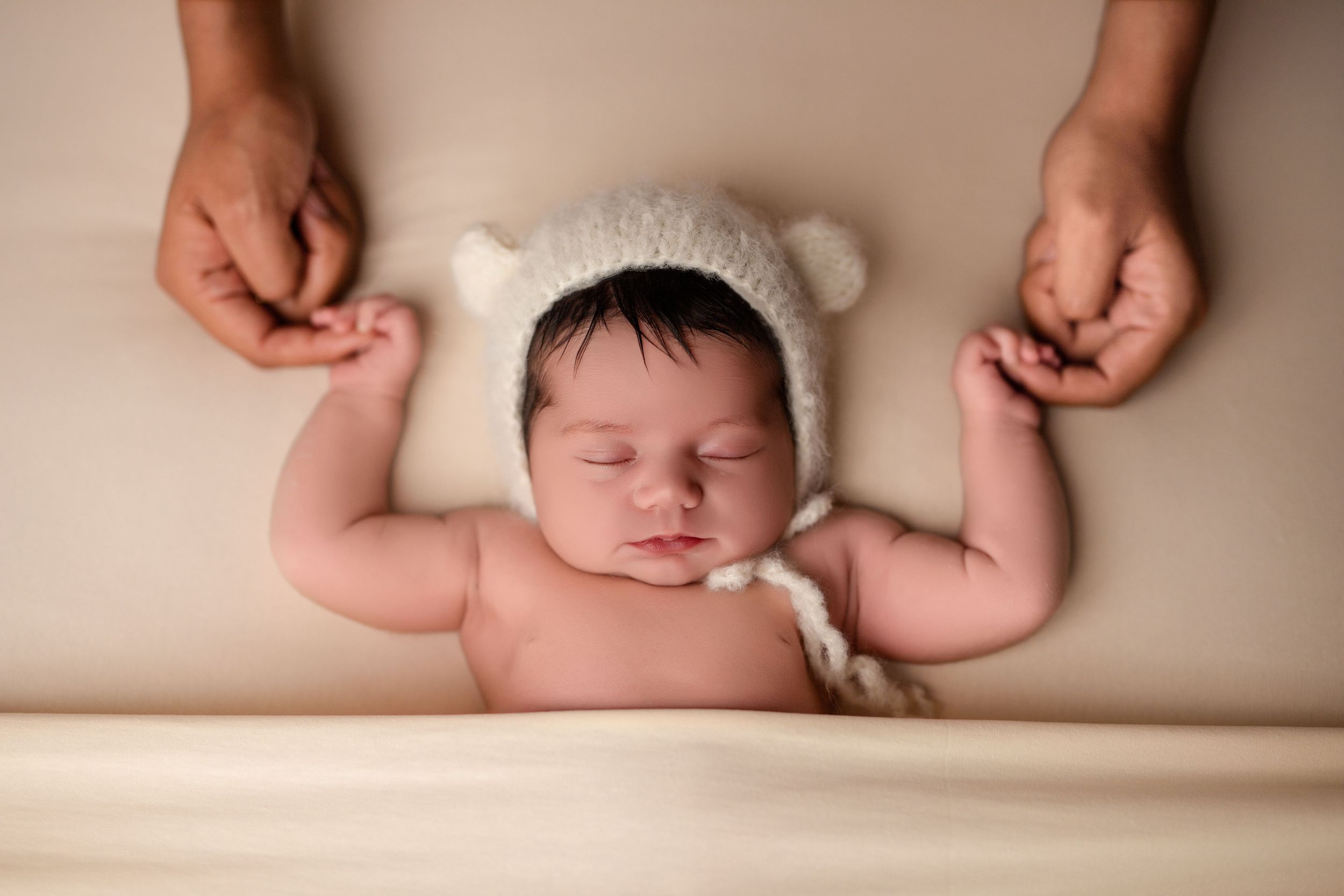 Sleeping newborn wearing white bear hat with hands gently held.