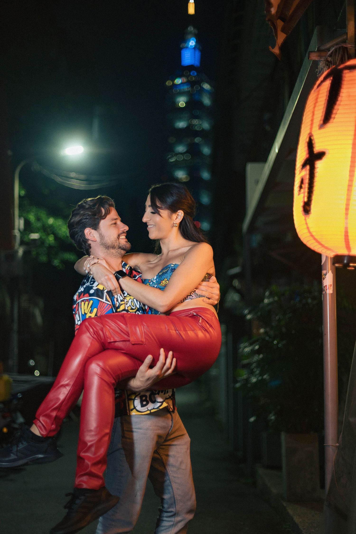 Man lifting woman in red pants while smiling together at night near Taipei 101 tower and paper lantern.