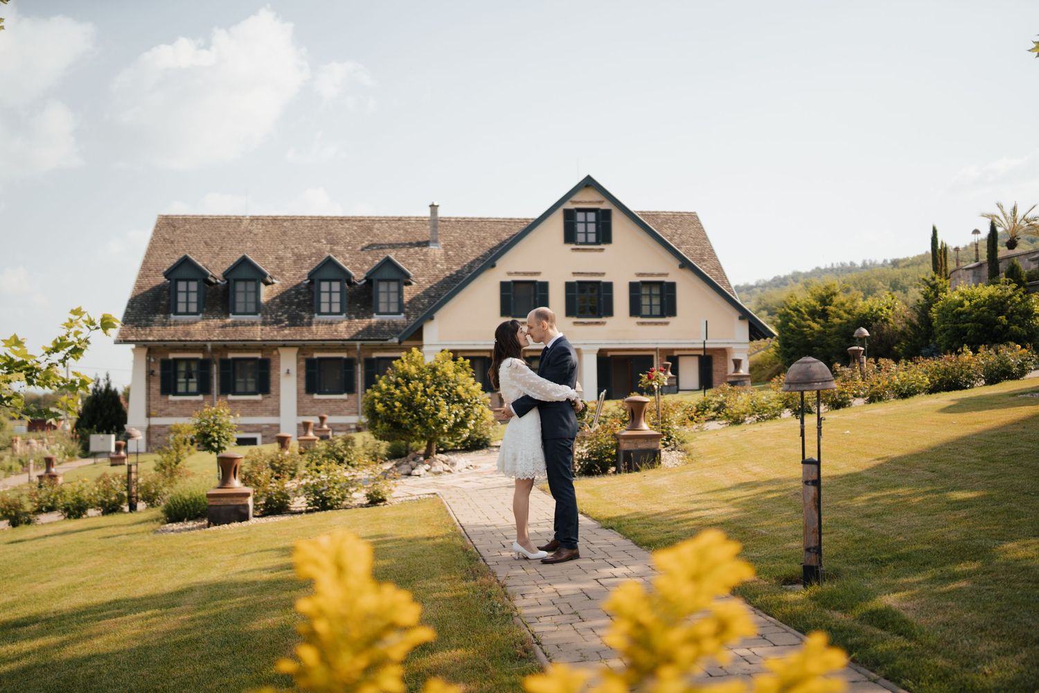A couple embraces on a garden path in front of a grand country house with yellow flowers in the foreground.