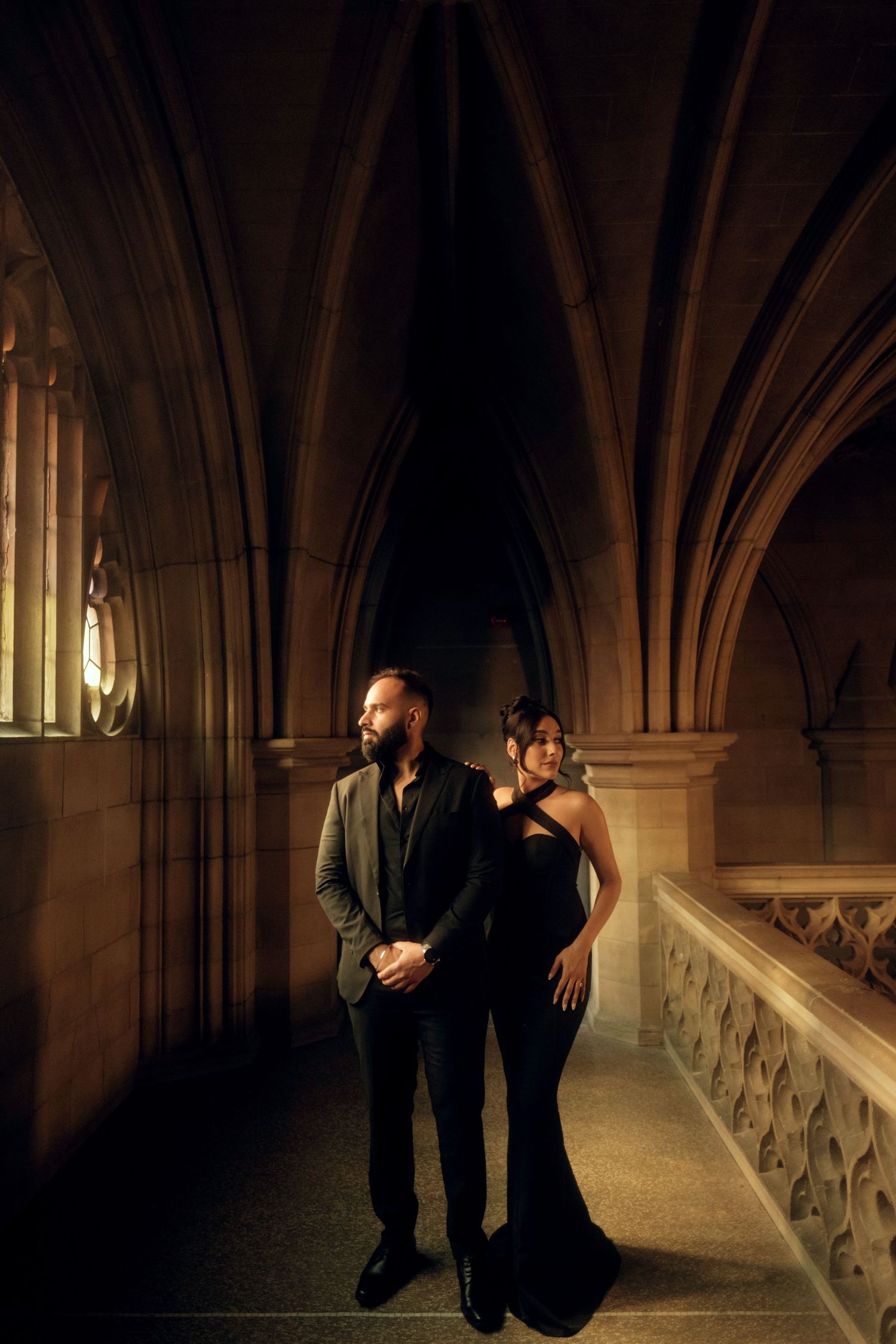 A couple in formal attire poses elegantly in a gothic cathedral hallway illuminated by warm lighting.