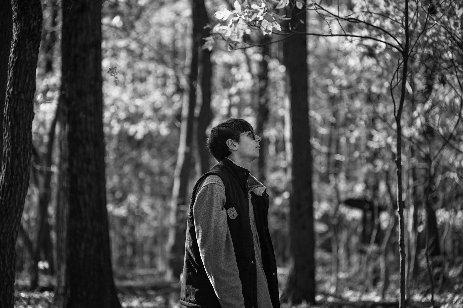 A black and white photo of a young man standing in a forest in autumn, with the light falling on the foliage of the trees behind him.