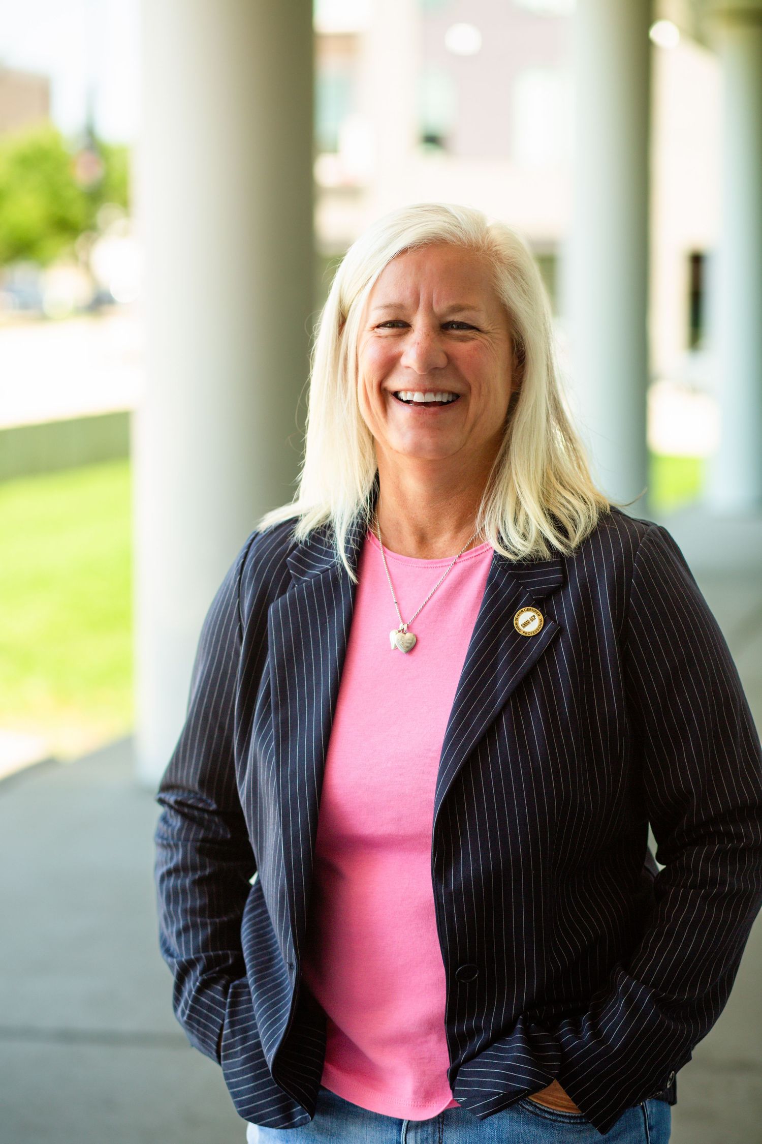 Portrait of someone in a navy pinstripe blazer and pink top standing in a covered outdoor walkway.