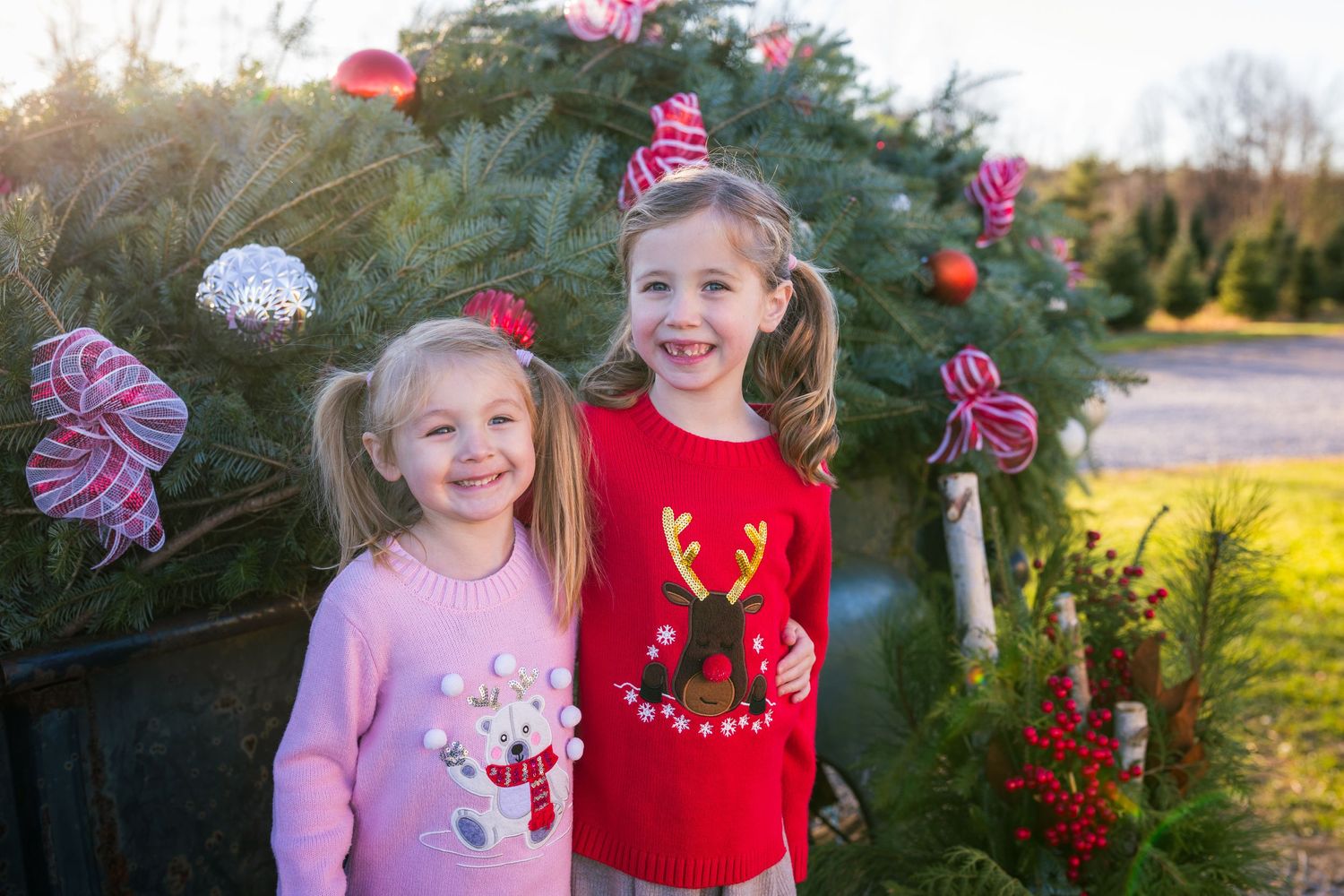 Two smiling children in festive sweaters posing by a decorated evergreen tree on a truck bed..