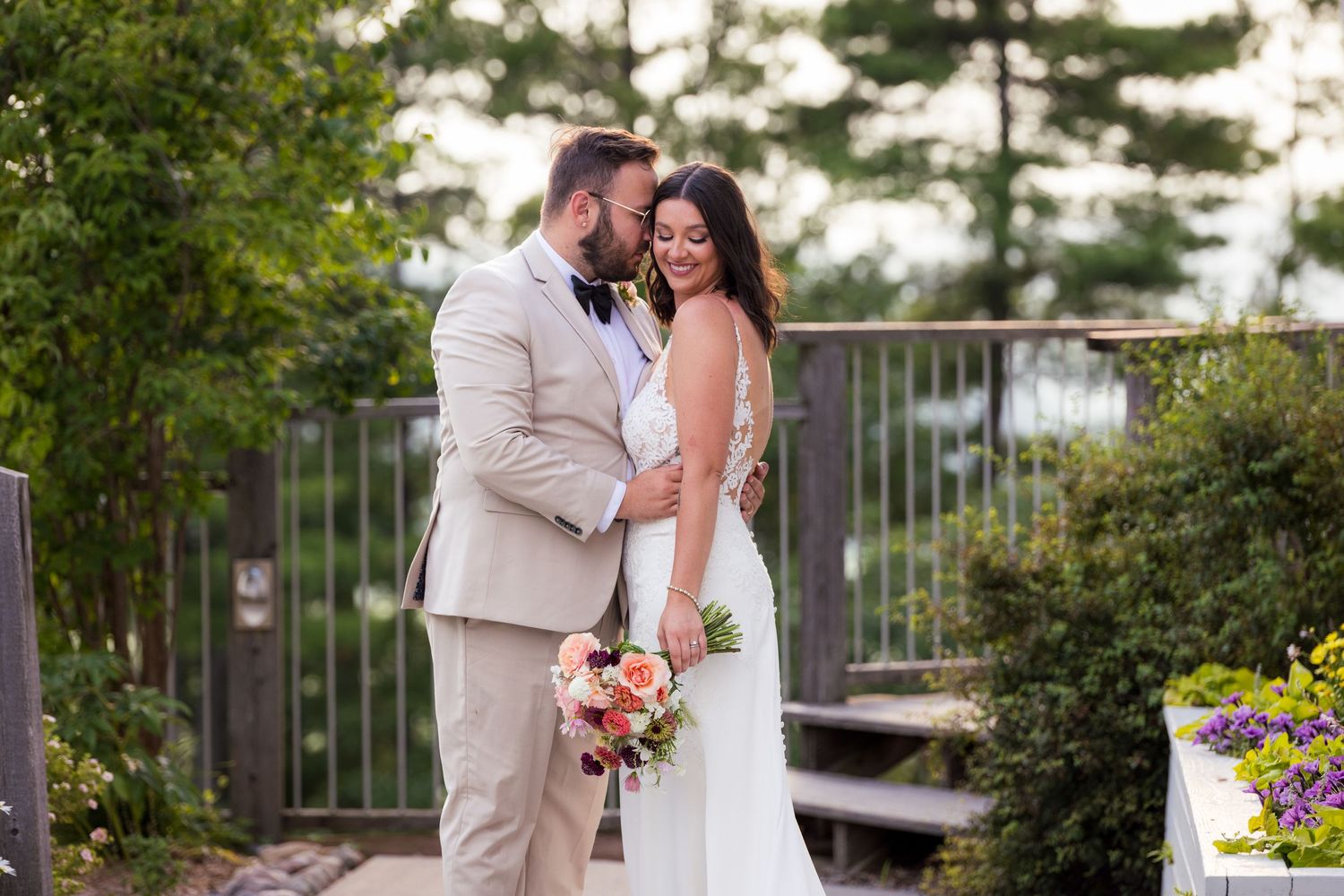 Groom in beige suit kisses bride in lace dress as she smiles, holding a colorful bouquet on a garden path with greenery around.