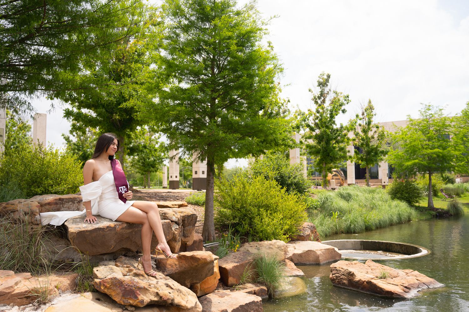 Person relaxes on stone ledge beside tranquil pond in landscaped garden with lush trees and greenery.