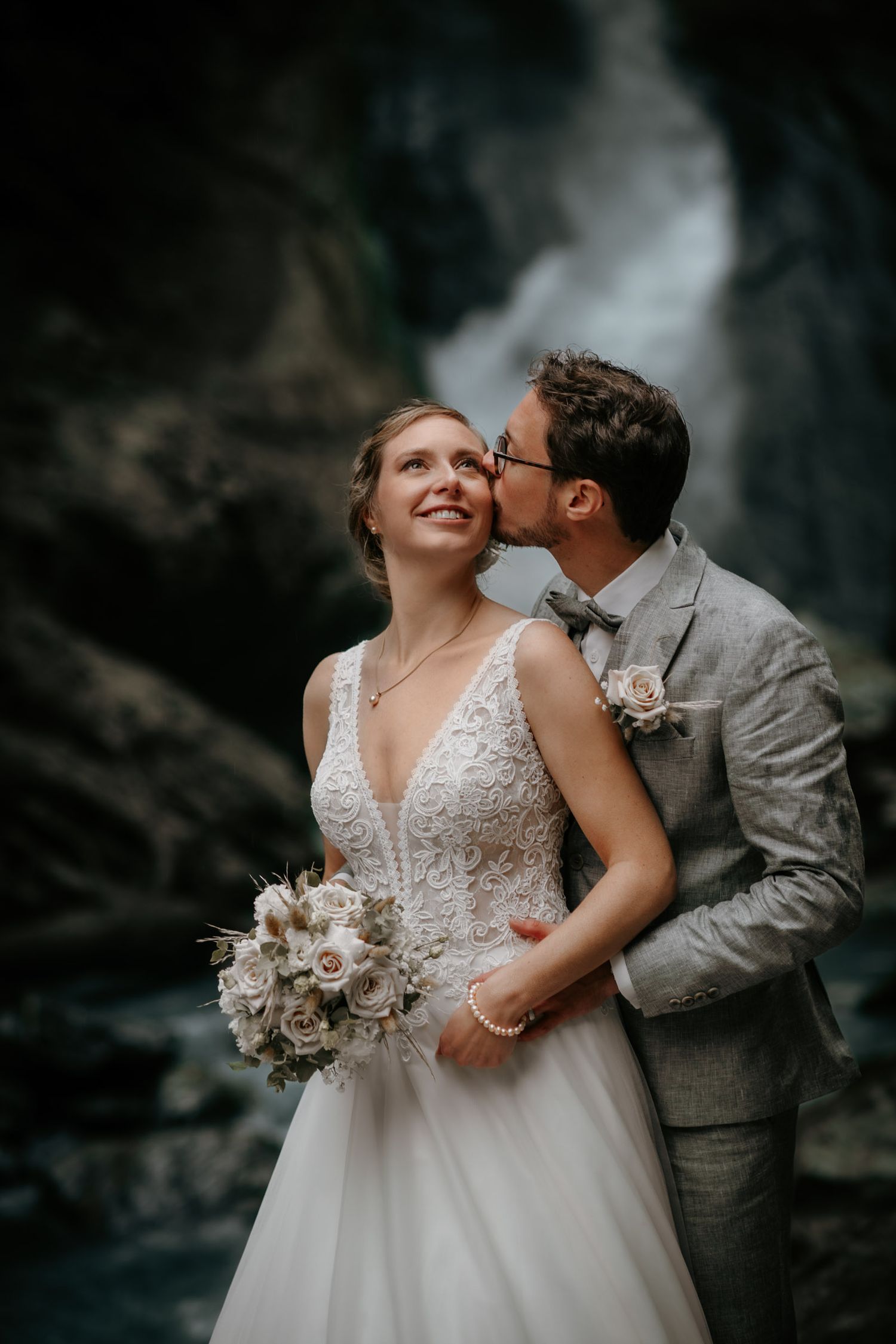 Groom in grey suit kissing bride on cheek before a dramatic waterfall backdrop.