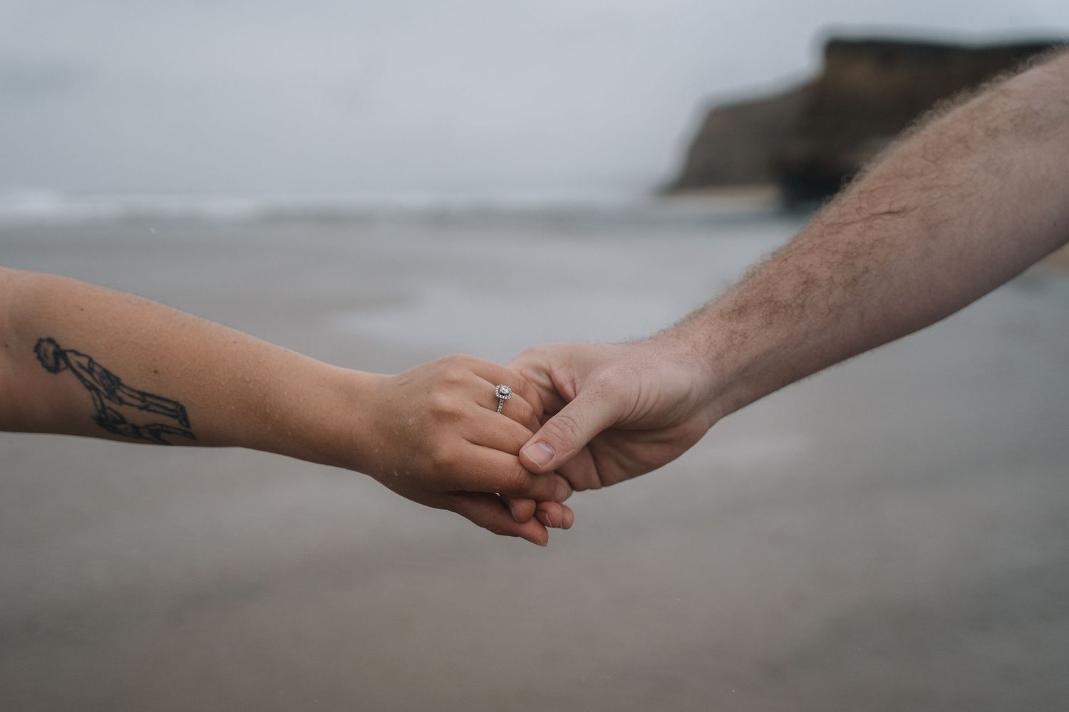 Two hands tenderly holding each other against a misty beach backdrop with small tattoo visible on one arm.