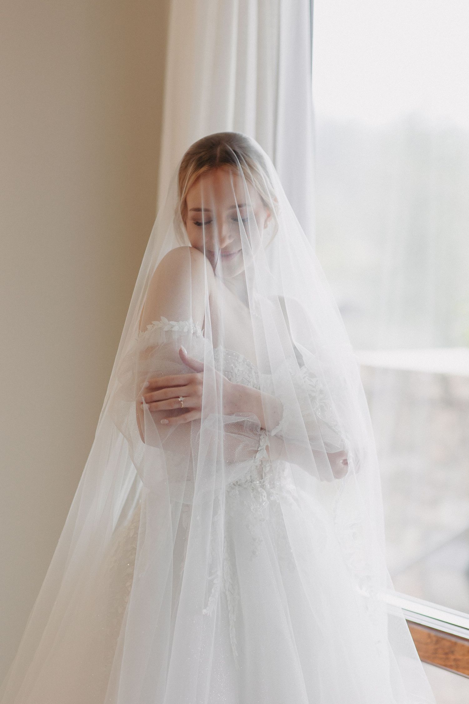 Ethereal bridal portrait through a soft white veil by a window.