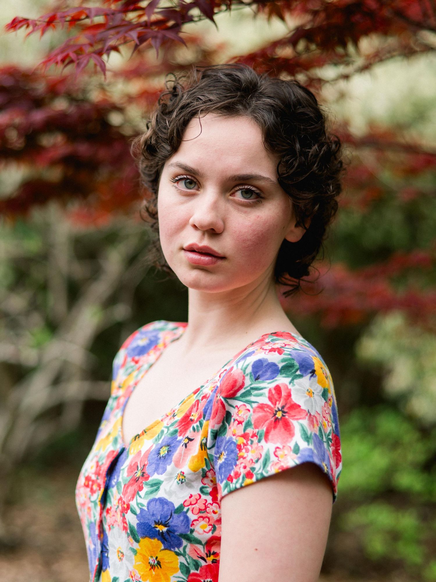 A floral print dress stands out against a backdrop of red Japanese maple leaves in an outdoor portrait.