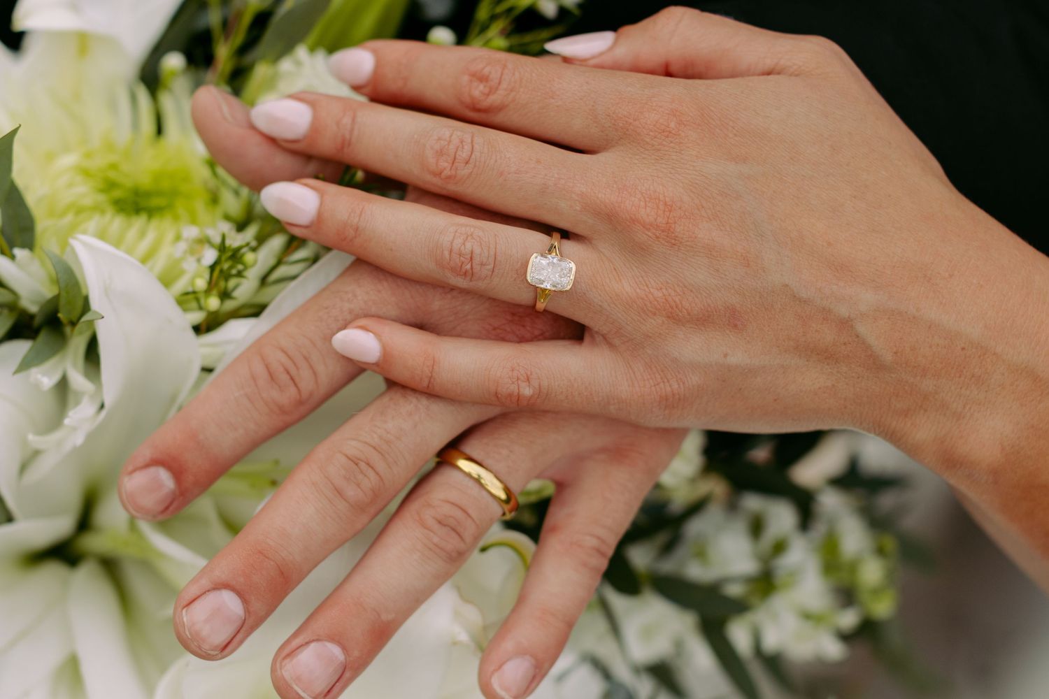 Two hands displaying wedding rings, one with a diamond engagement ring and one with a gold band, resting on white flowers.