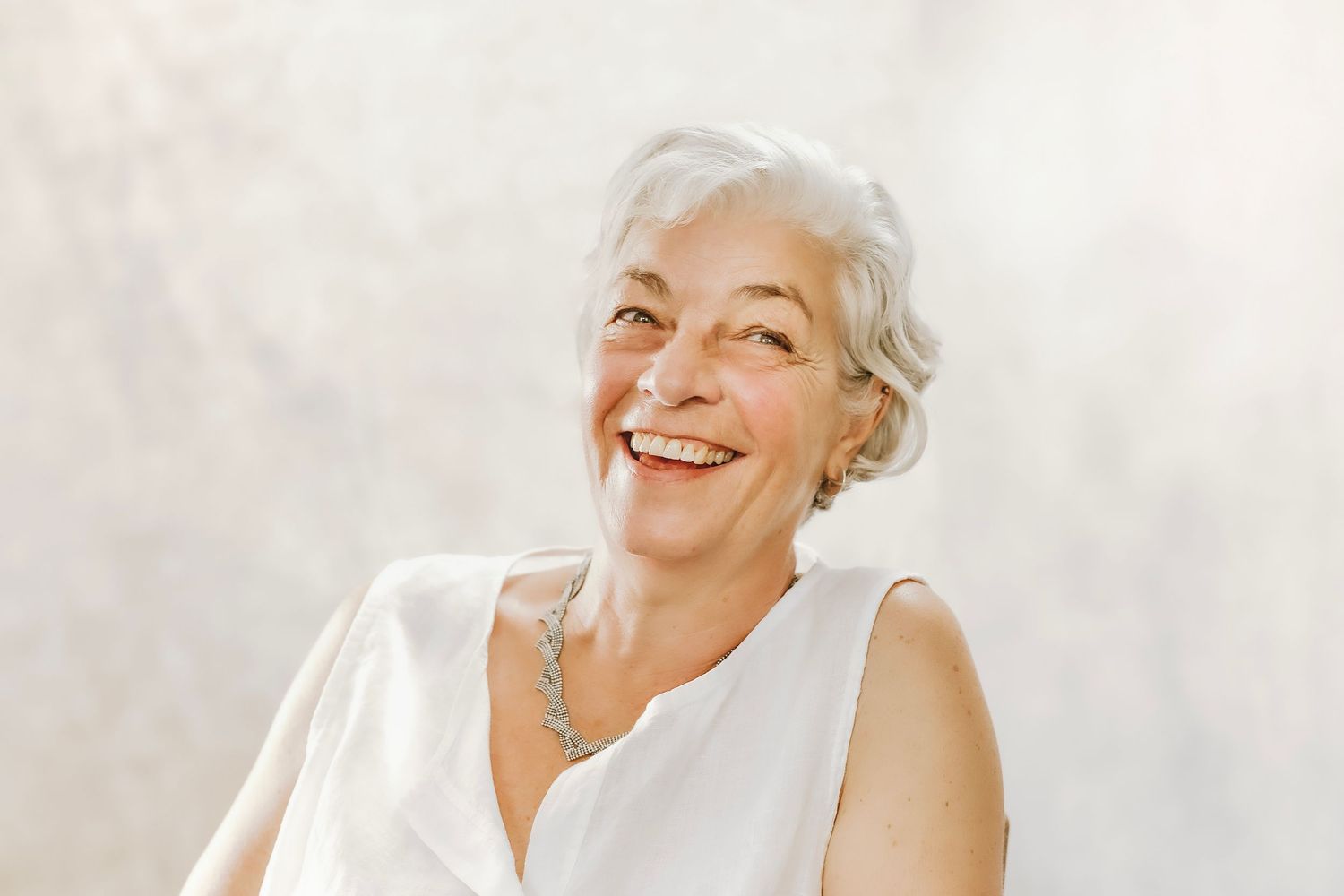 Smiling older woman with short gray hair wearing a white sleeveless top and silver necklace against a light background.
