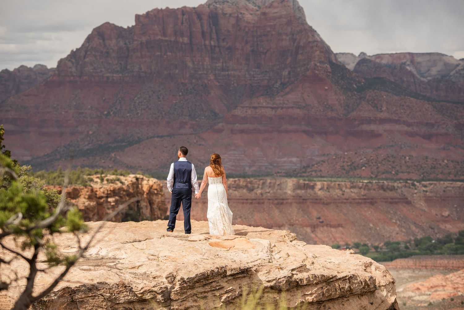 Couple in wedding attire standing on a rocky cliff overlooking red rock canyon landscape at Zion National Park.