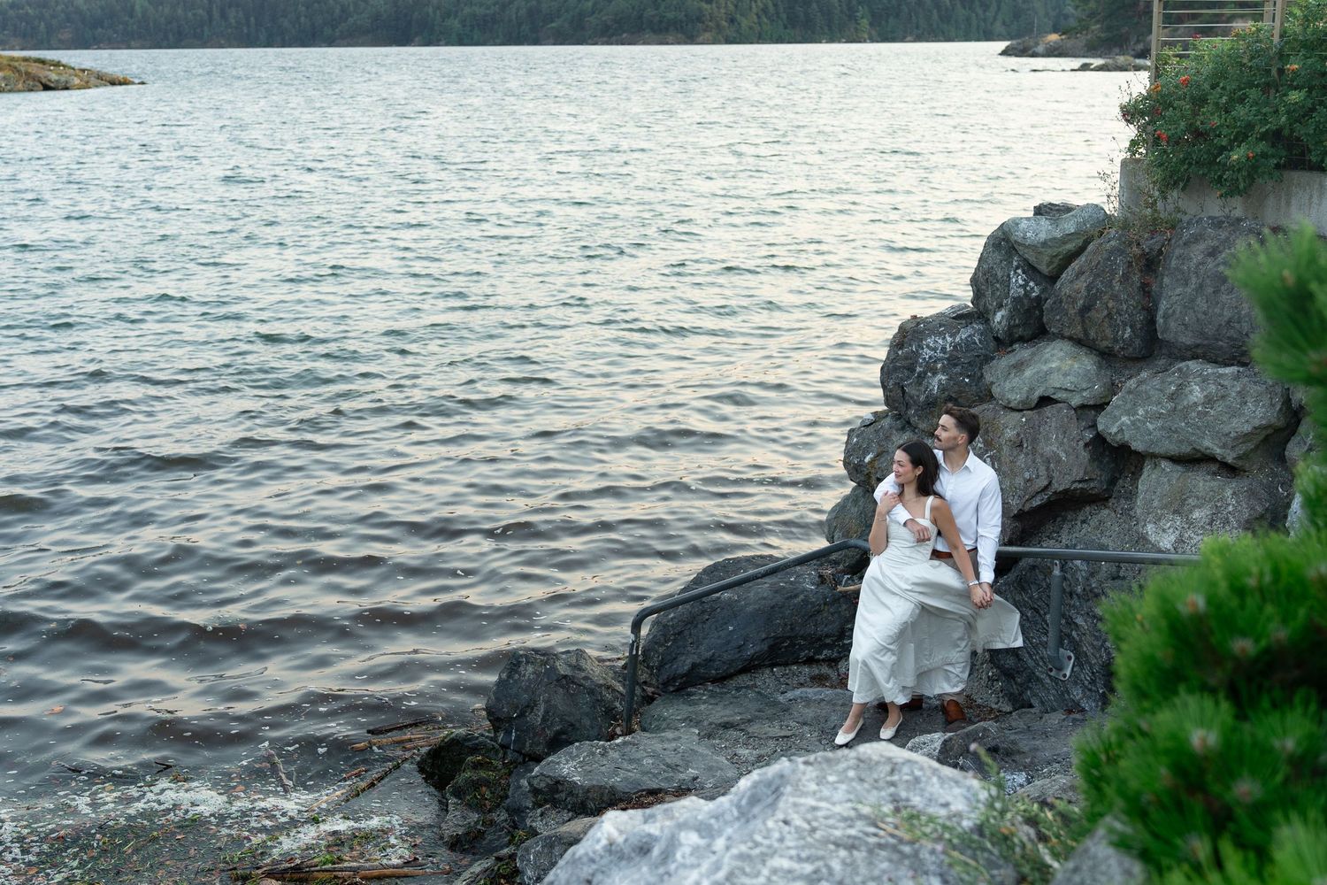 Couple in white sitting on rocky shoreline by calm lake surrounded by lush trees at dusk.