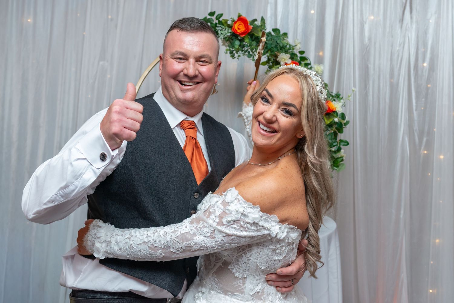 A smiling bride in lace gown and floral crown poses with groom in vest and orange tie under a greenery arch.