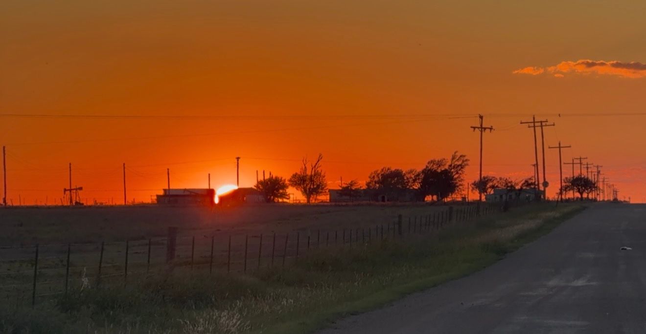 Vibrant orange sunset over rural farmland with silhouetted trees, power lines, and buildings along a quiet country road.