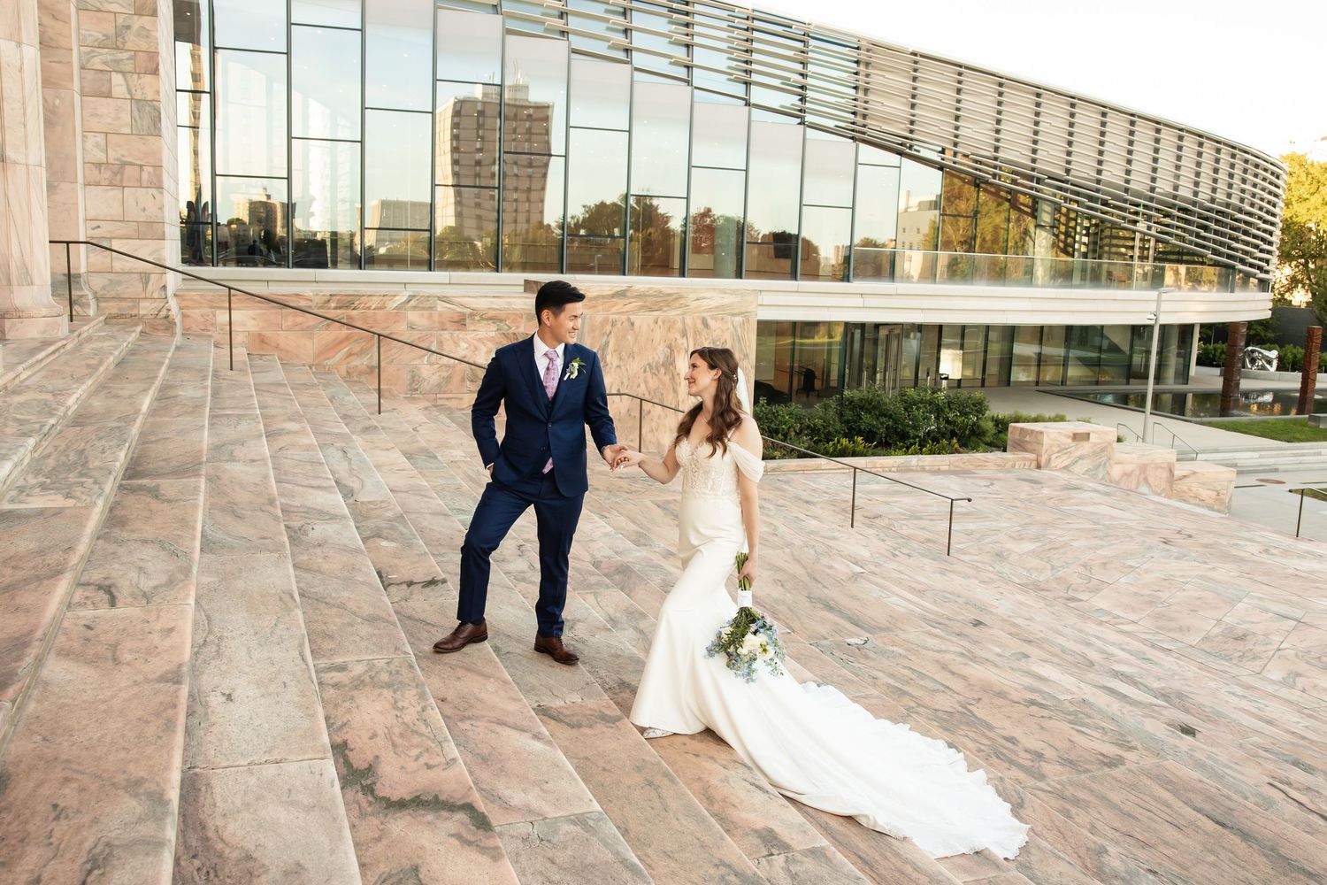 Bride and groom walk hand-in-hand on stone steps outside a modern glass building during their wedding day.