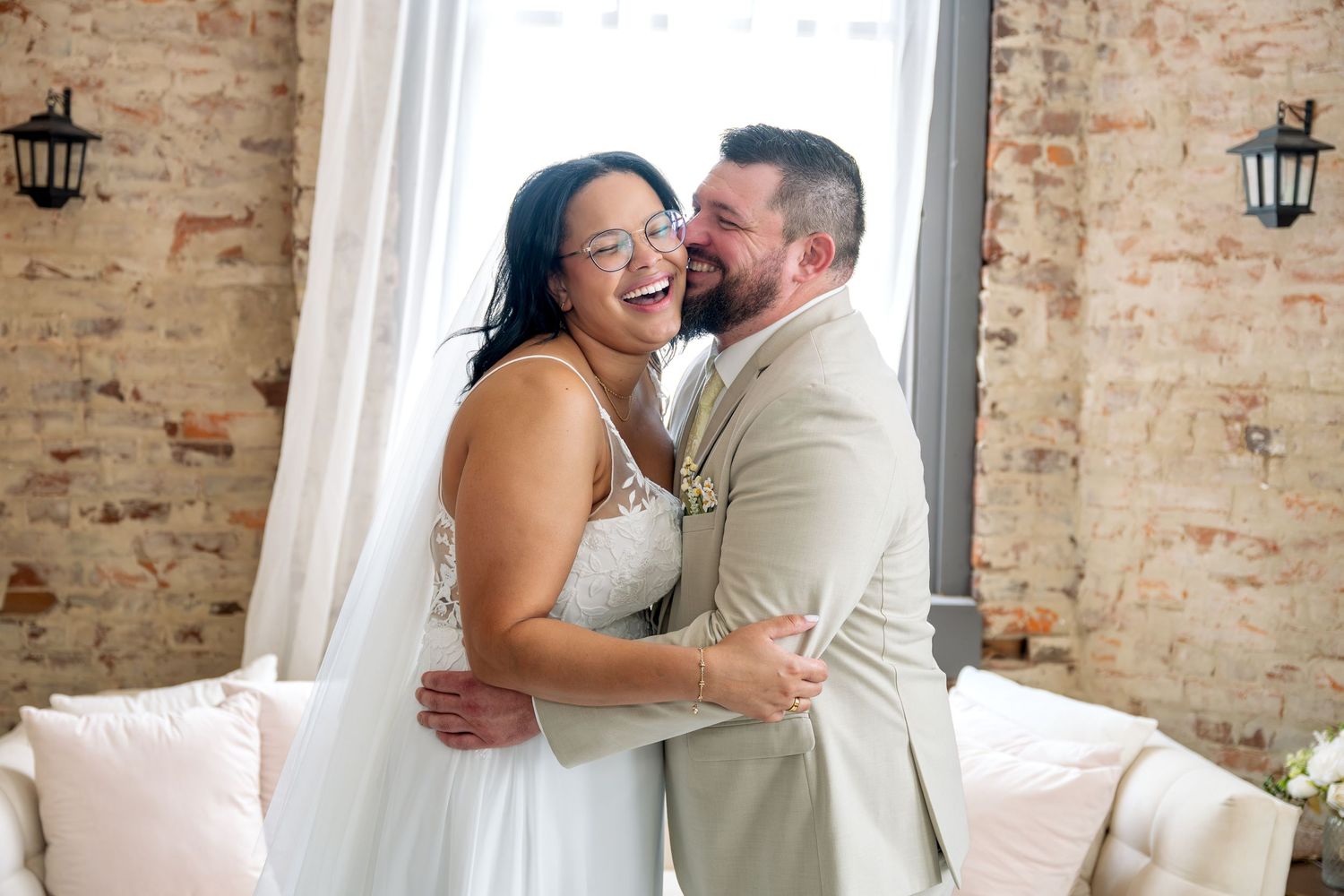 A joyful bride and groom share a laughing embrace in a rustic brick-walled venue on their wedding day.