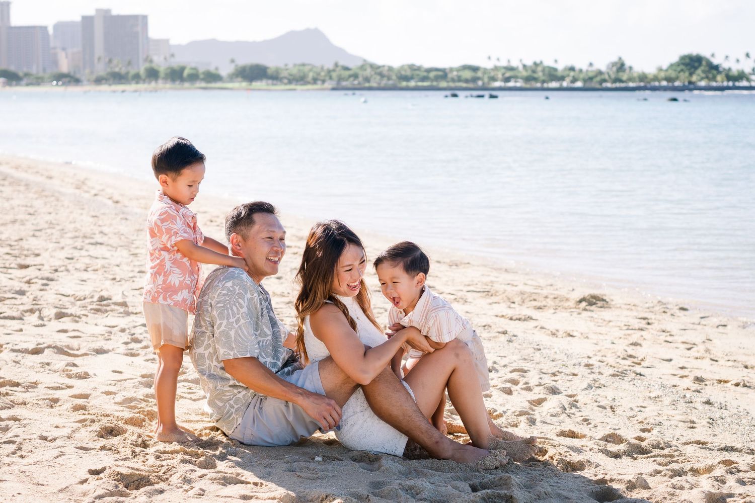 Happy family of four sitting on sandy beach with ocean and Diamond Head crater in background on sunny day.