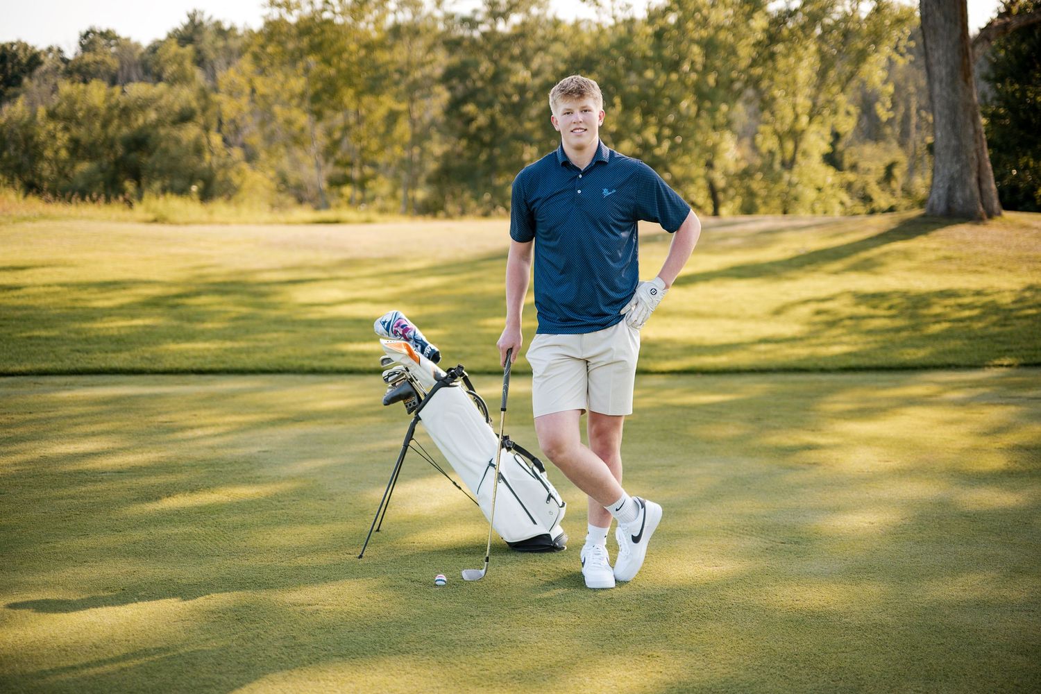 Senior guy golfer in navy polo and khaki shorts stands beside a white golf bag on green course at Blackwolf Run for senior photos