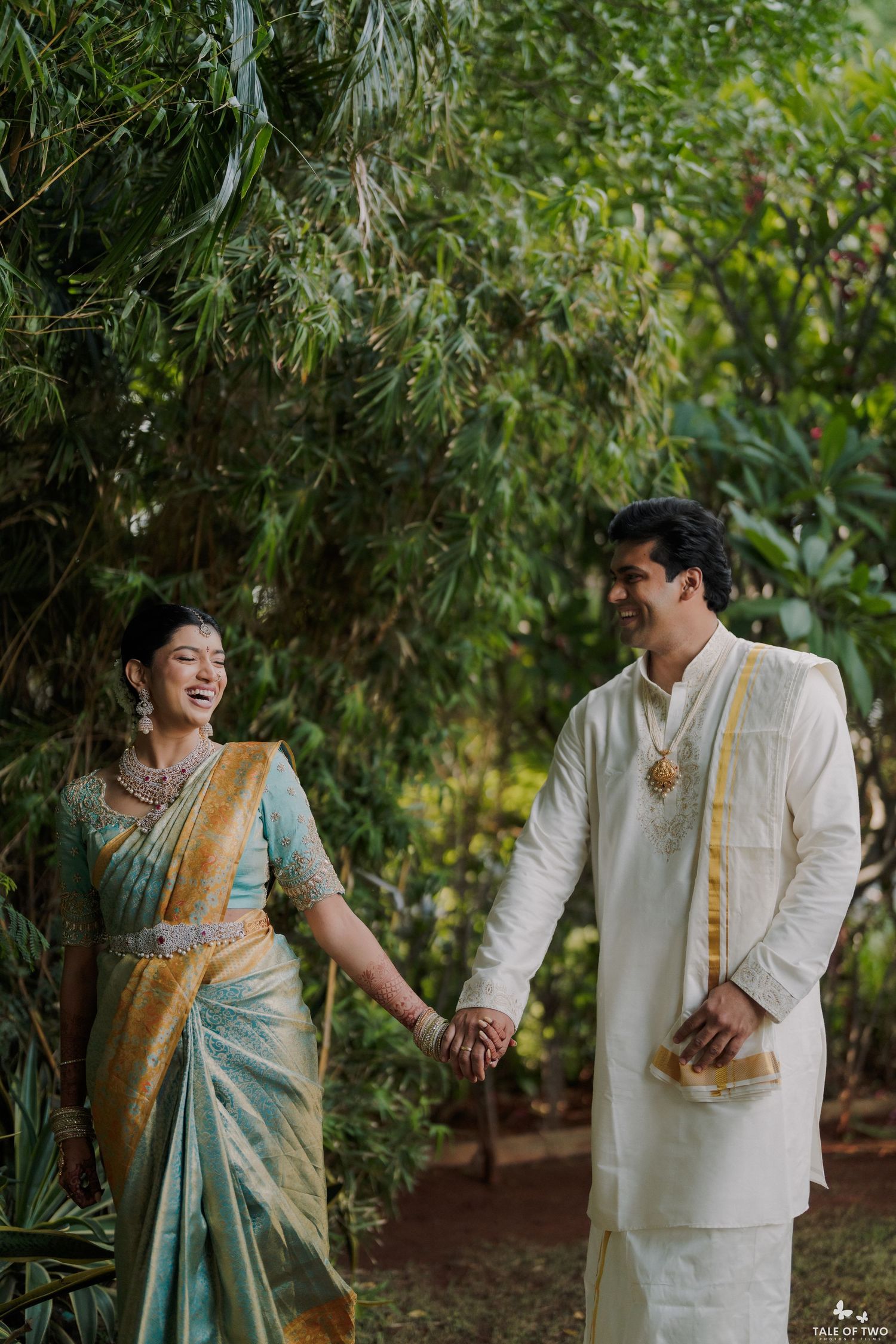 Indian couple in traditional wedding attire holding hands outdoors, bride in teal saree, groom in white sherwani.