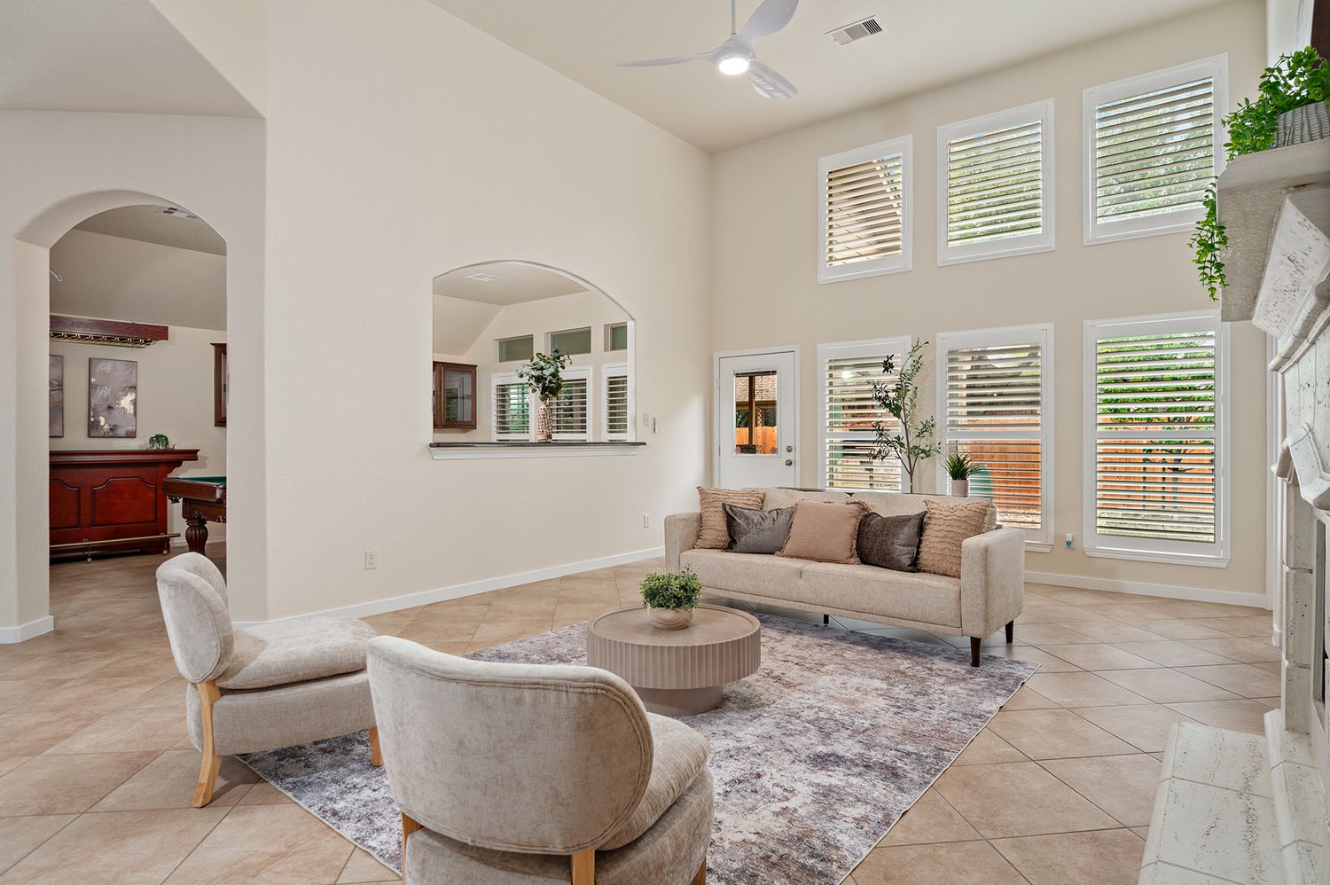 Bright living room with tile floors, plush neutral sofa, cushioned armchairs, round coffee table, plantation shutters and an arched opening.