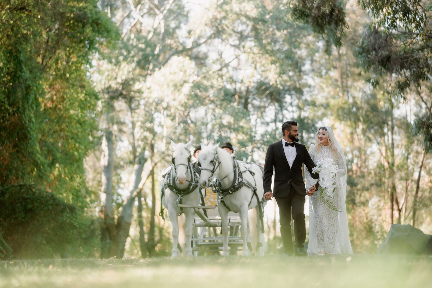 Bride in a lace gown and veil holding a white floral bouquet stands beside a groom in a black tuxedo and bow tie among tall forest trees.