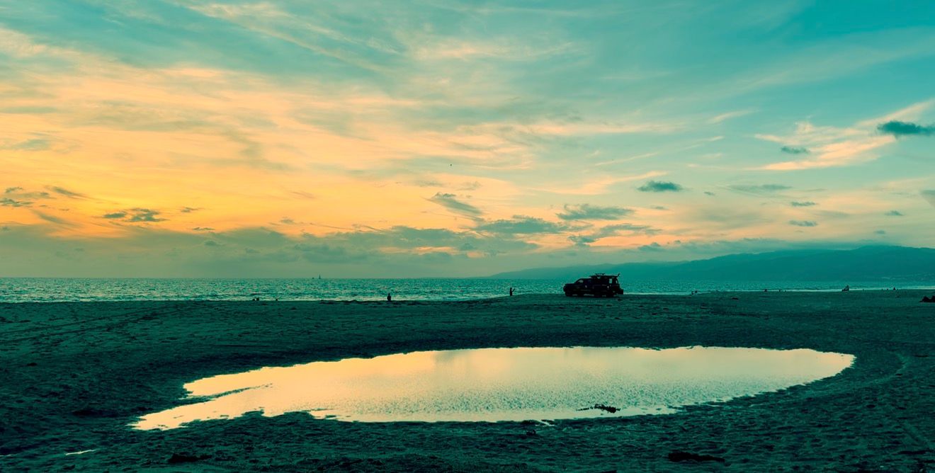 A tidal pool reflects golden sunset light on a wide sandy beach under a colorful sky with soft clouds and distant hazy mountains.