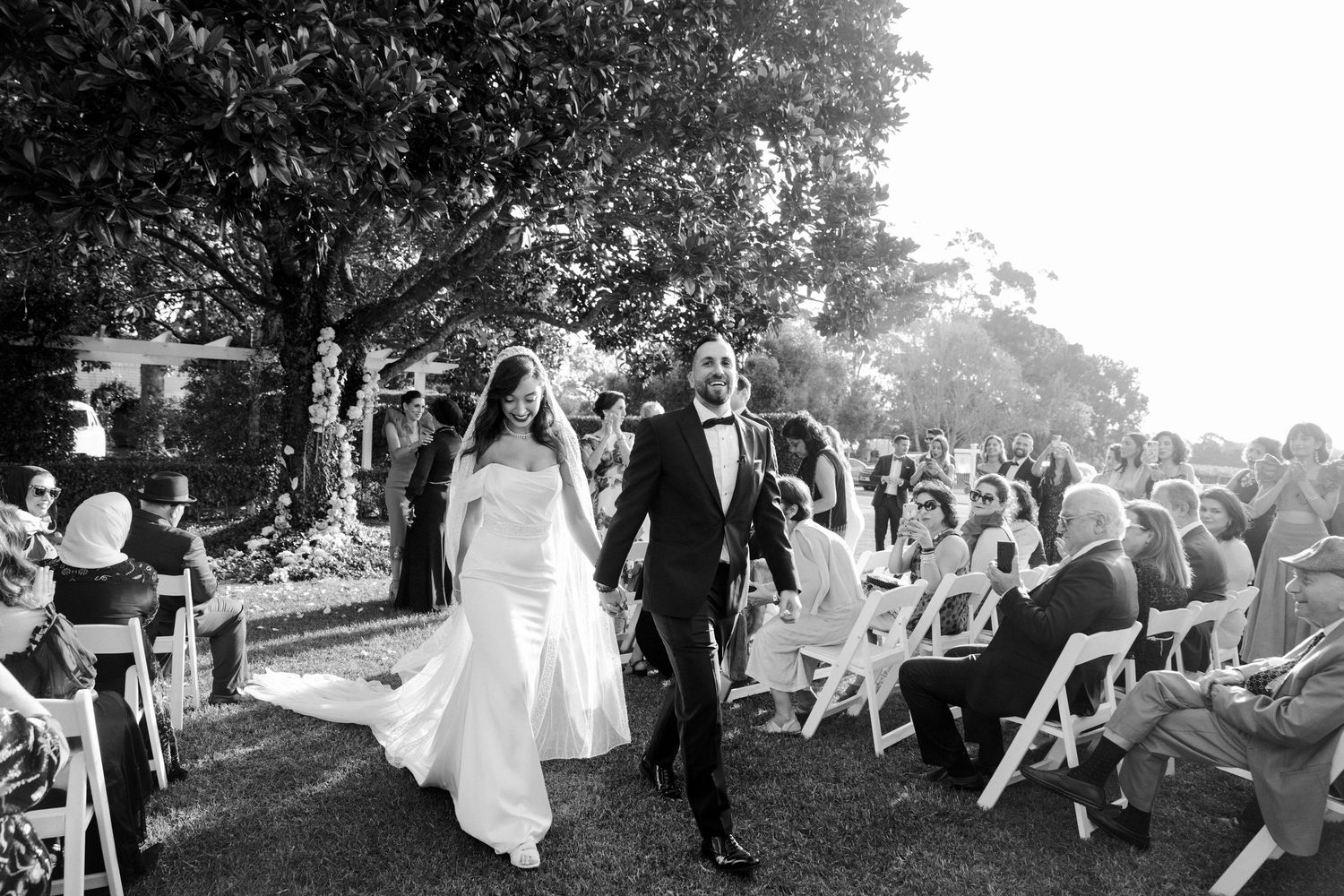 Black and white photo of newlyweds walking down outdoor aisle after wedding ceremony, bride in gown with veil, groom in tuxedo.