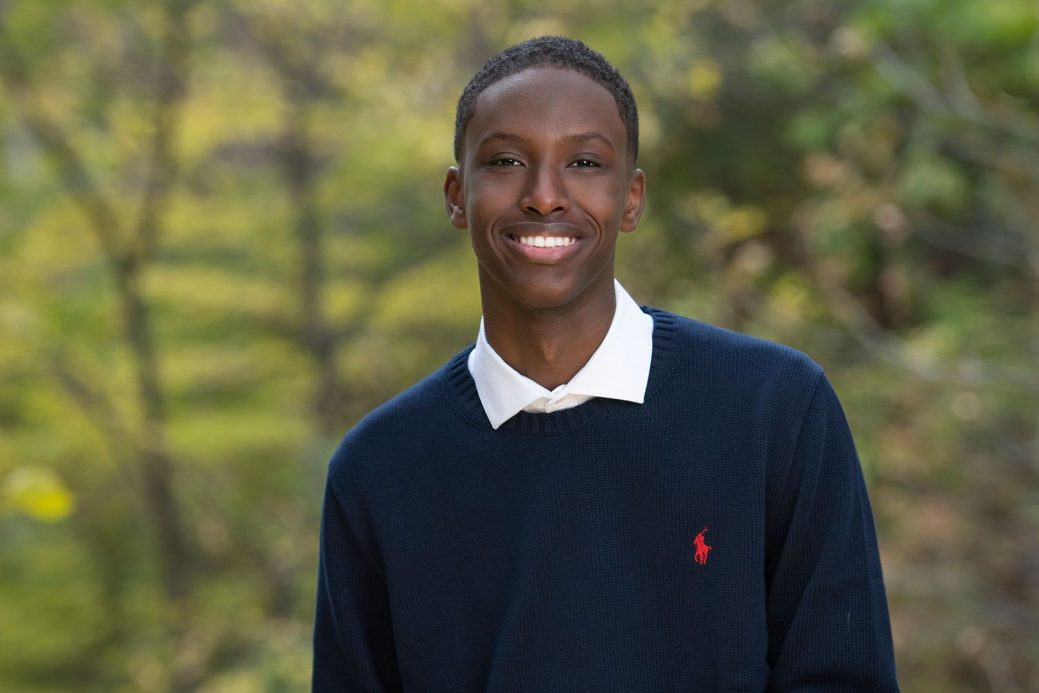 Smiling young man wearing a navy Polo Ralph Lauren sweater outdoors with green foliage background.
