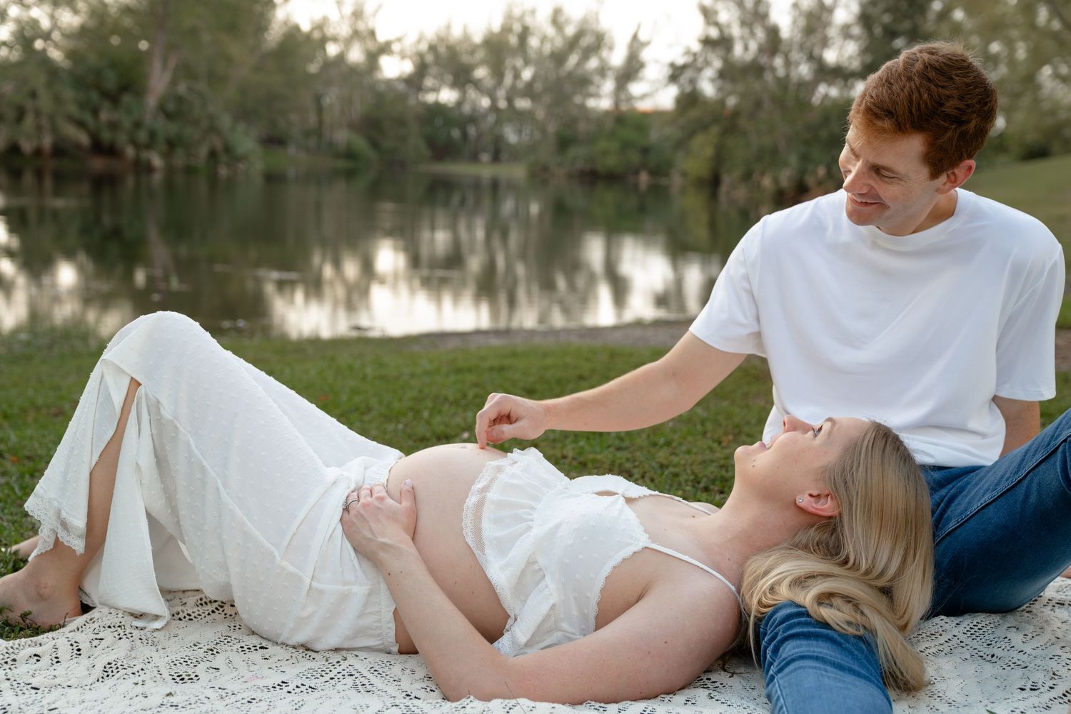 Pregnant couple relaxing by a lake, woman lying down as man sits beside her touching her baby bump.