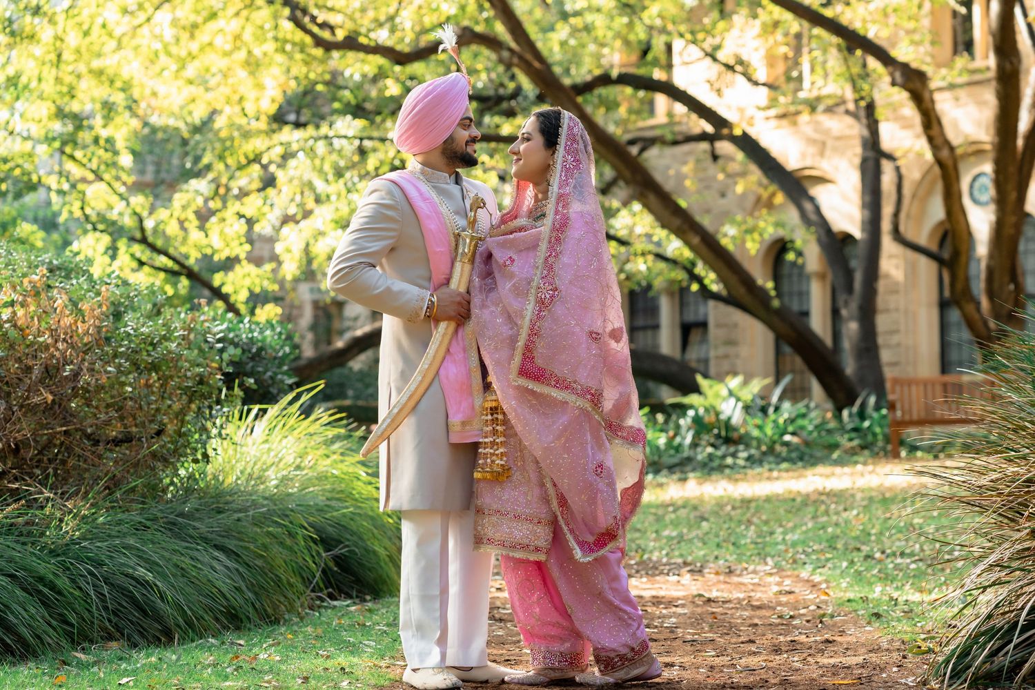Sikh couple in traditional pink and gold wedding attire embrace outdoors among lush green trees.