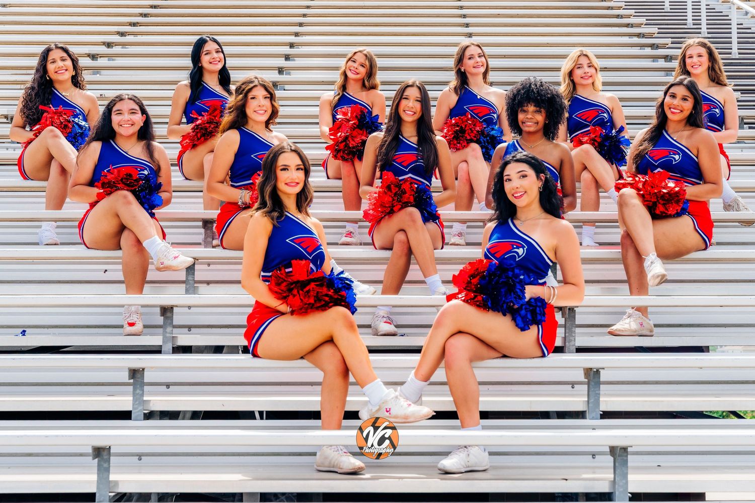 Cheerleaders in red and blue uniforms pose on bleachers holding pom-poms at a football stadium.