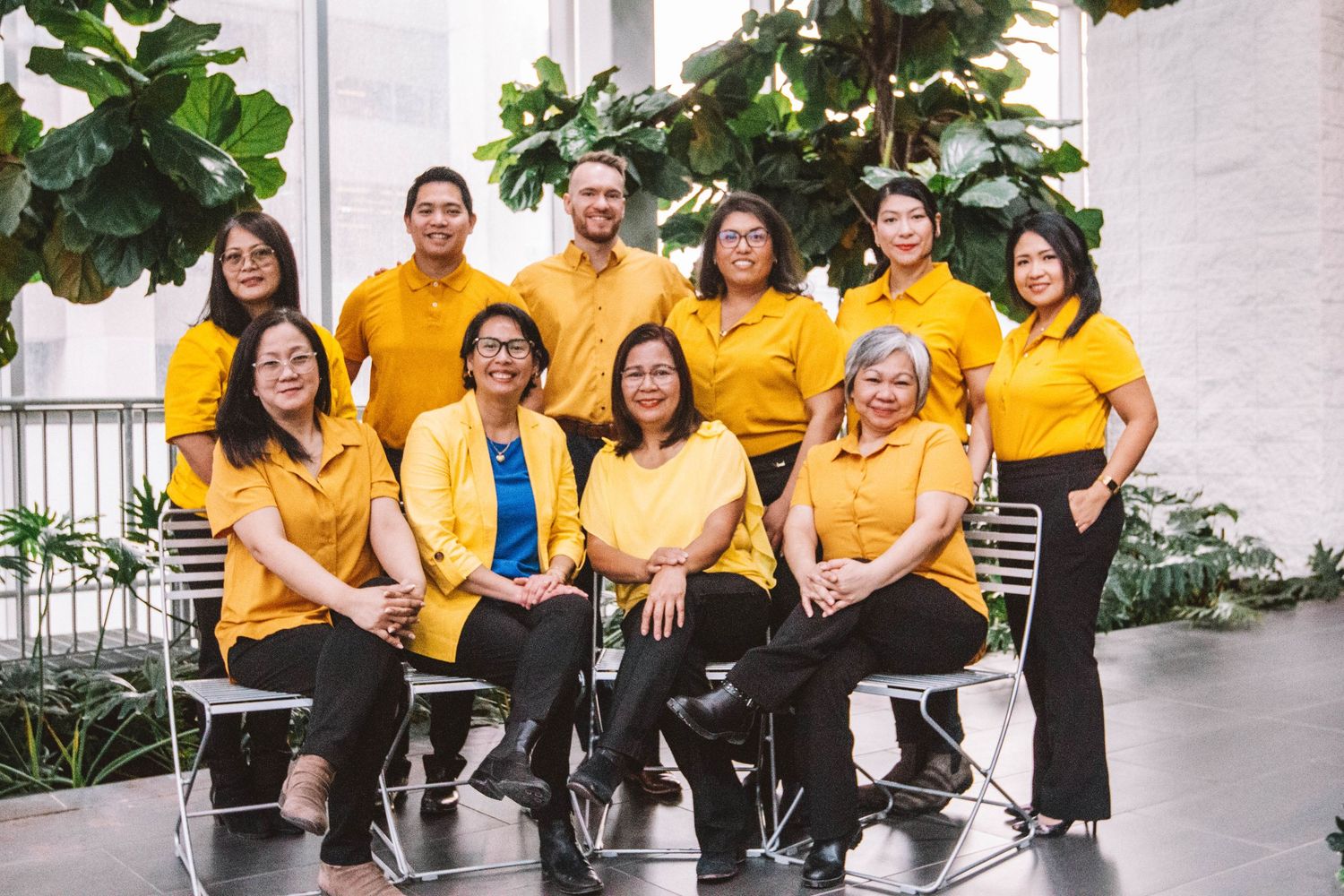 Group of ten professionals in matching yellow polo shirts posing together indoors with tropical plants in background.