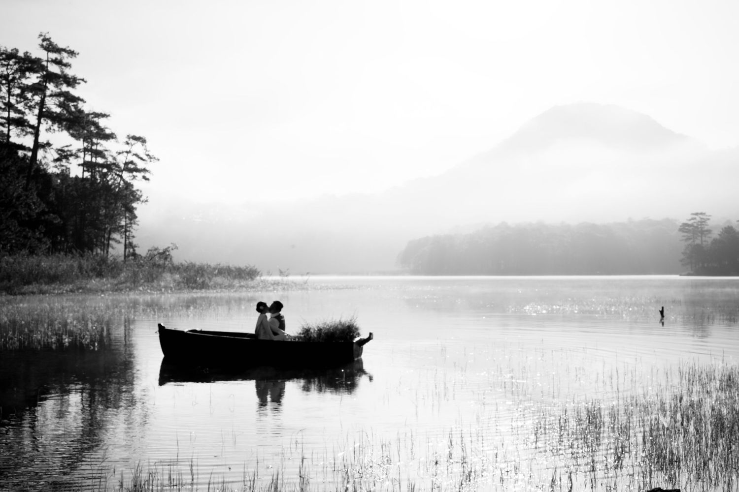 Black and white photo of a couple sitting in a rowboat on a calm misty lake with mountains and trees in background.