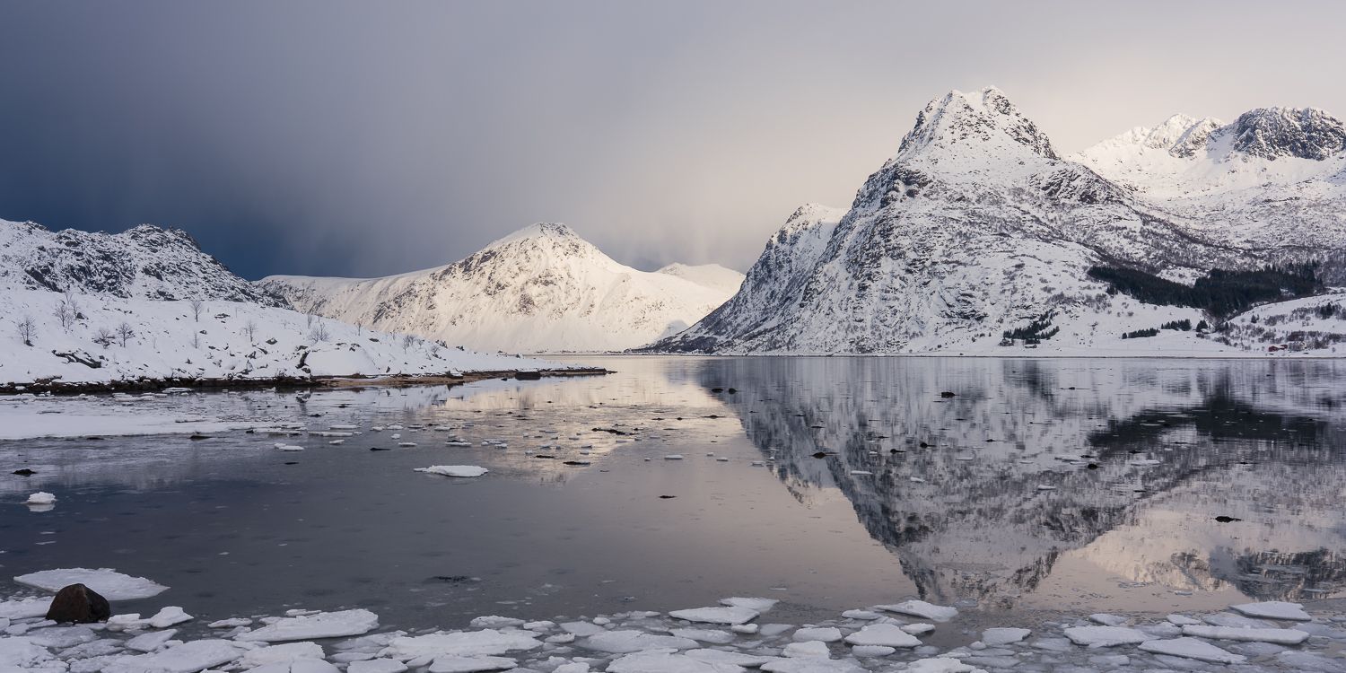 Snow-covered mountains reflecting in a partially frozen fjord under dramatic winter storm clouds in Norway.
