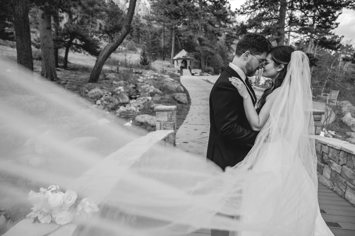 Bride and groom walk hand in hand along a paved path surrounded by tall pine trees at an outdoor wedding venue.
