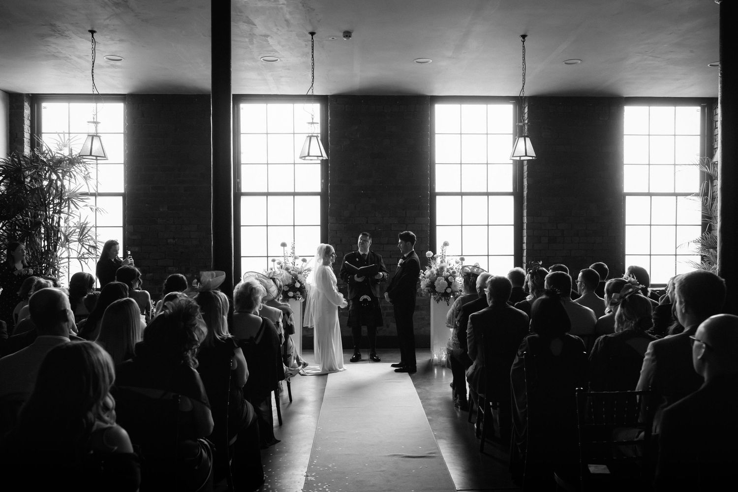 Bride and groom pose in front of a glass greenhouse, she in a strapless gown, he in a black tuxedo.