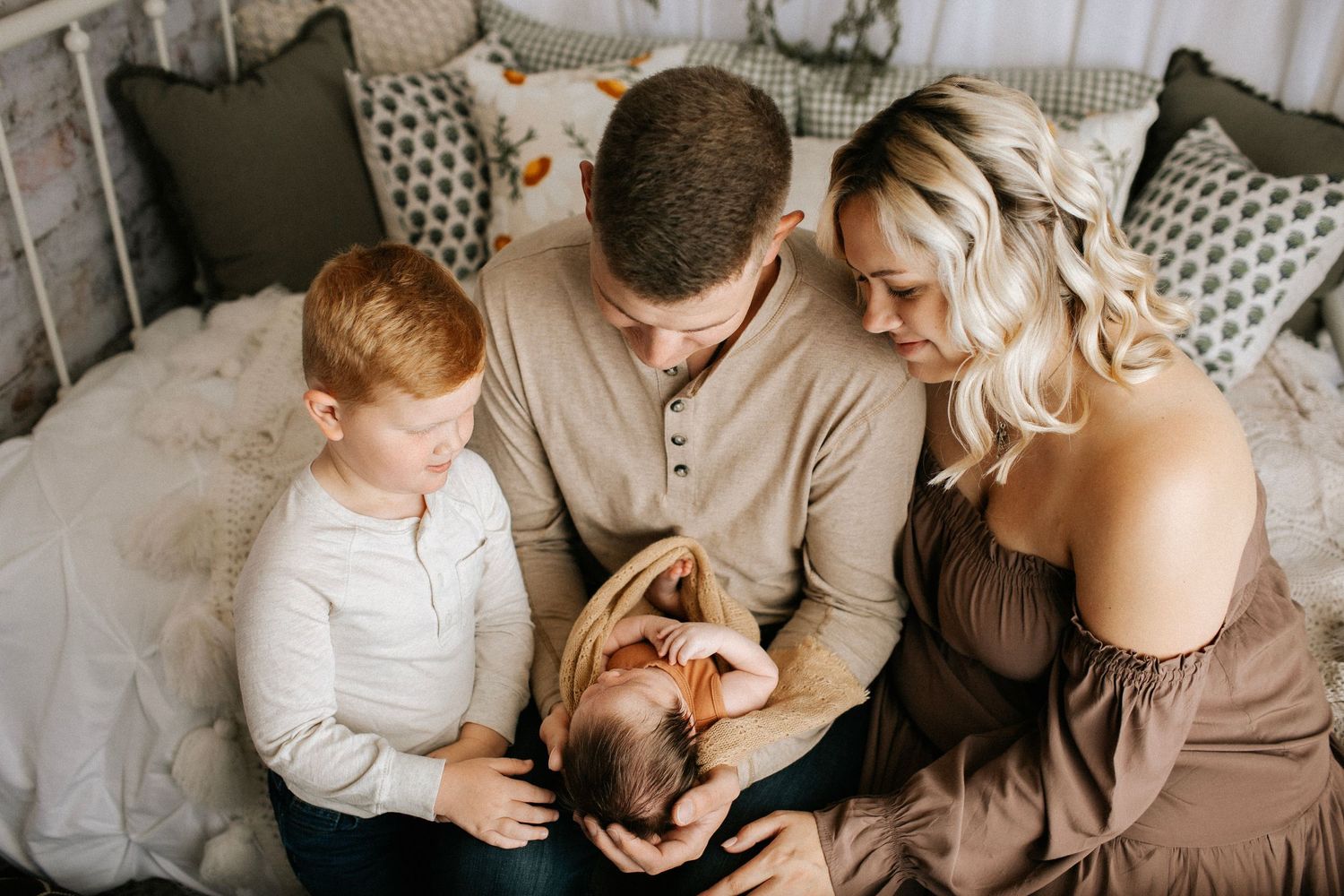 Family of four admiring their newborn baby together, seated on a bed in a cozy bedroom setting.