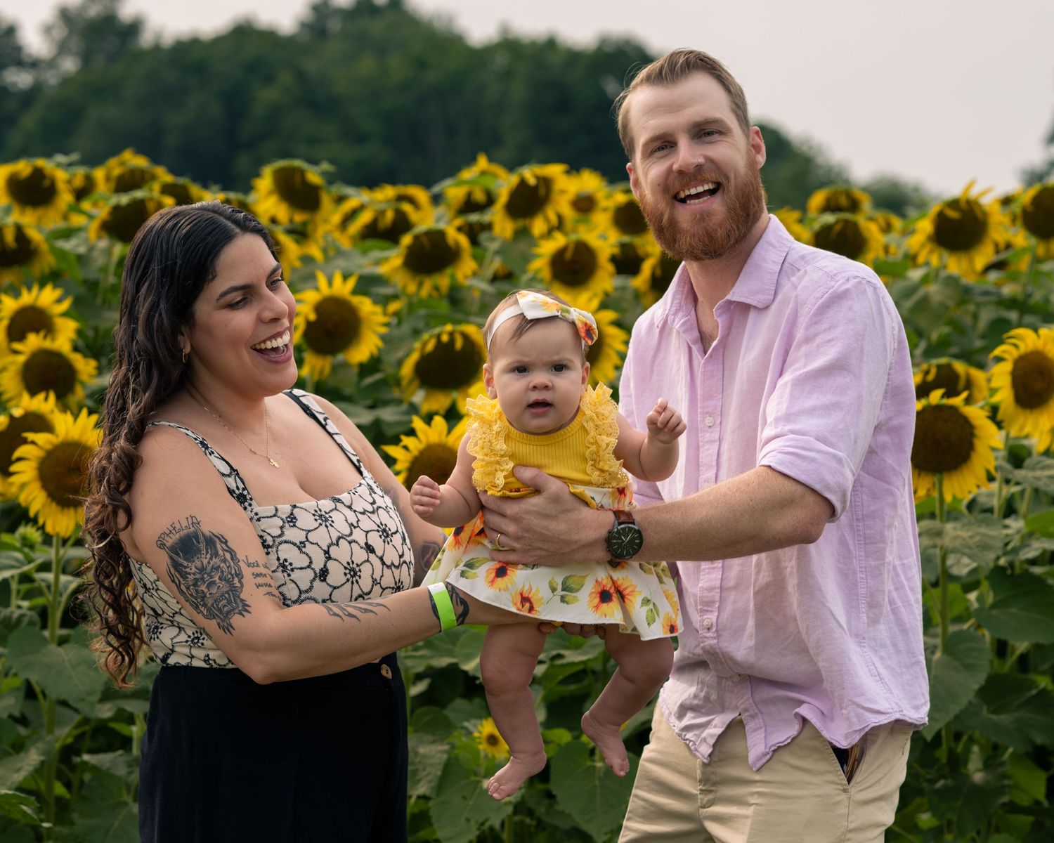 Happy family with tattooed mom, bearded dad holding baby girl in yellow dress, posing in sunny sunflower field.