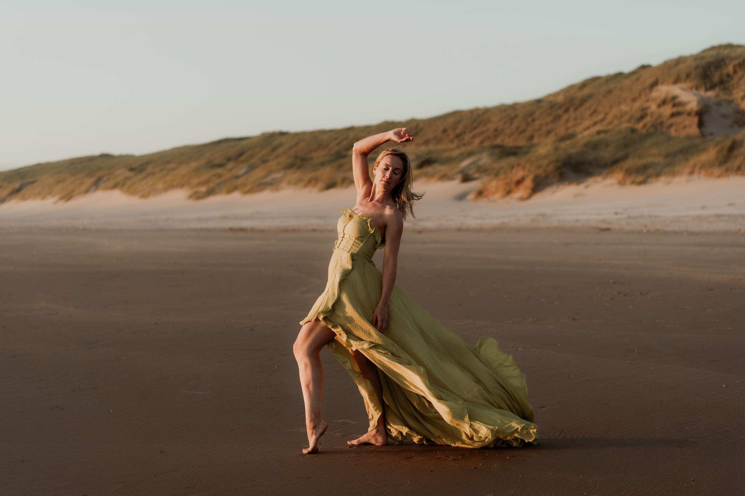 Woman in flowing green gown poses gracefully on a sandy beach with dunes in the background.