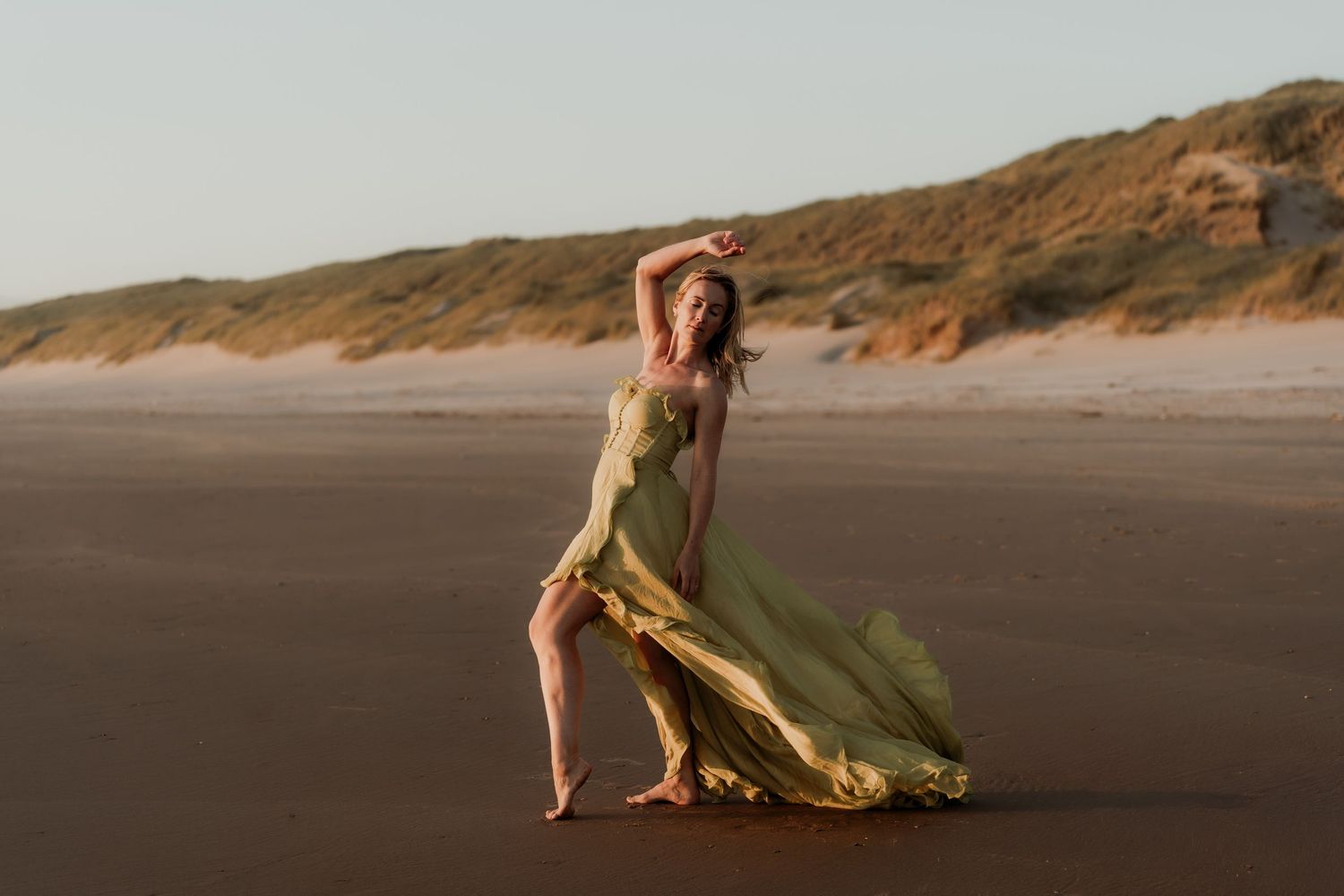 Woman in flowing green gown poses gracefully on a sandy beach with dunes in the background.